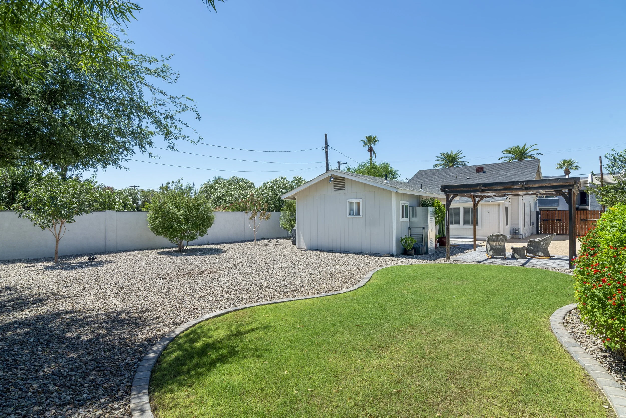 Resilient backyard with lush green lawn, landscaped garden, mature trees, and a small white shed with seating area under a pergola, all enclosed by a white privacy fence with a clear blue sky overhead.