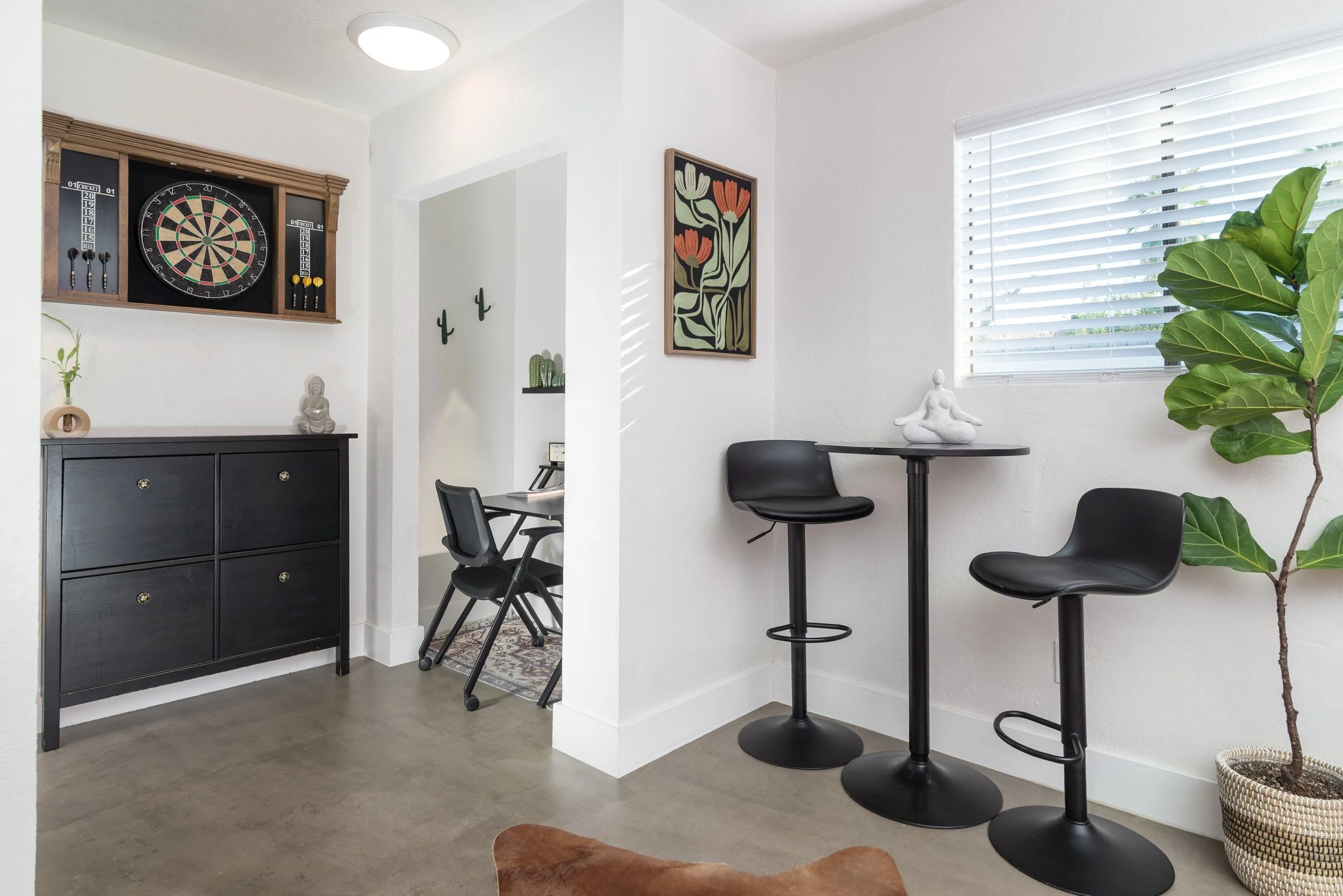 A cozy indoor space featuring a black bar-height table with two matching stools, a large leafy green plant, a white sculpture on the table, a framed floral painting on the wall, and a window with white blinds allowing natural light. There is also a dark wooden cabinet under a dartboard and some decorative cactus plants on a shelf.