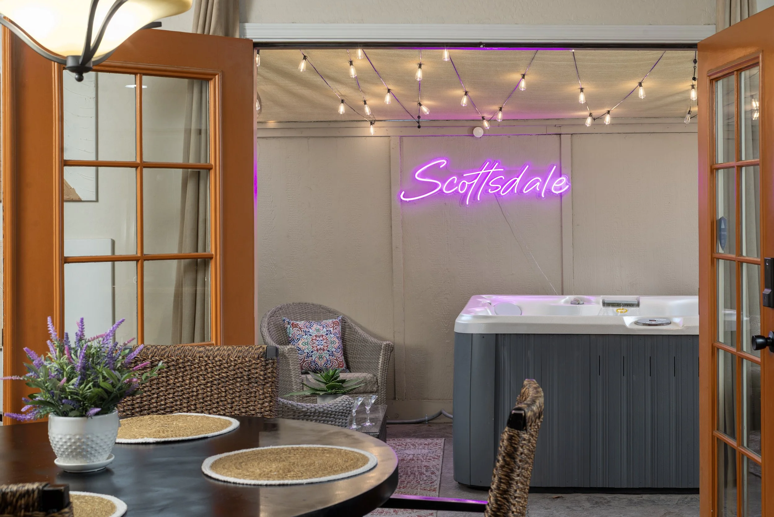 Open French doors leading into a patio or indoor space with a neon pink "Scottsdale" sign on the wall, string lights hanging from the ceiling, a wicker chair with a patterned pillow, a hot tub, and a table with a plant and woven placemats.