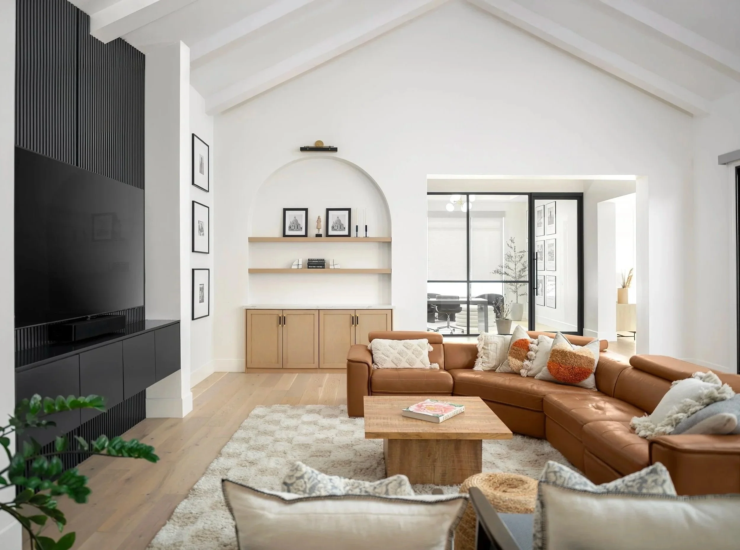 Modern living room with a large brown sectional sofa, wooden coffee table, beige and white textured pillows, light hardwood floors, black entertainment center with a TV, built-in white shelves with decorations, glass sliding door separating an adjacent room, lights, and minimalist decor.