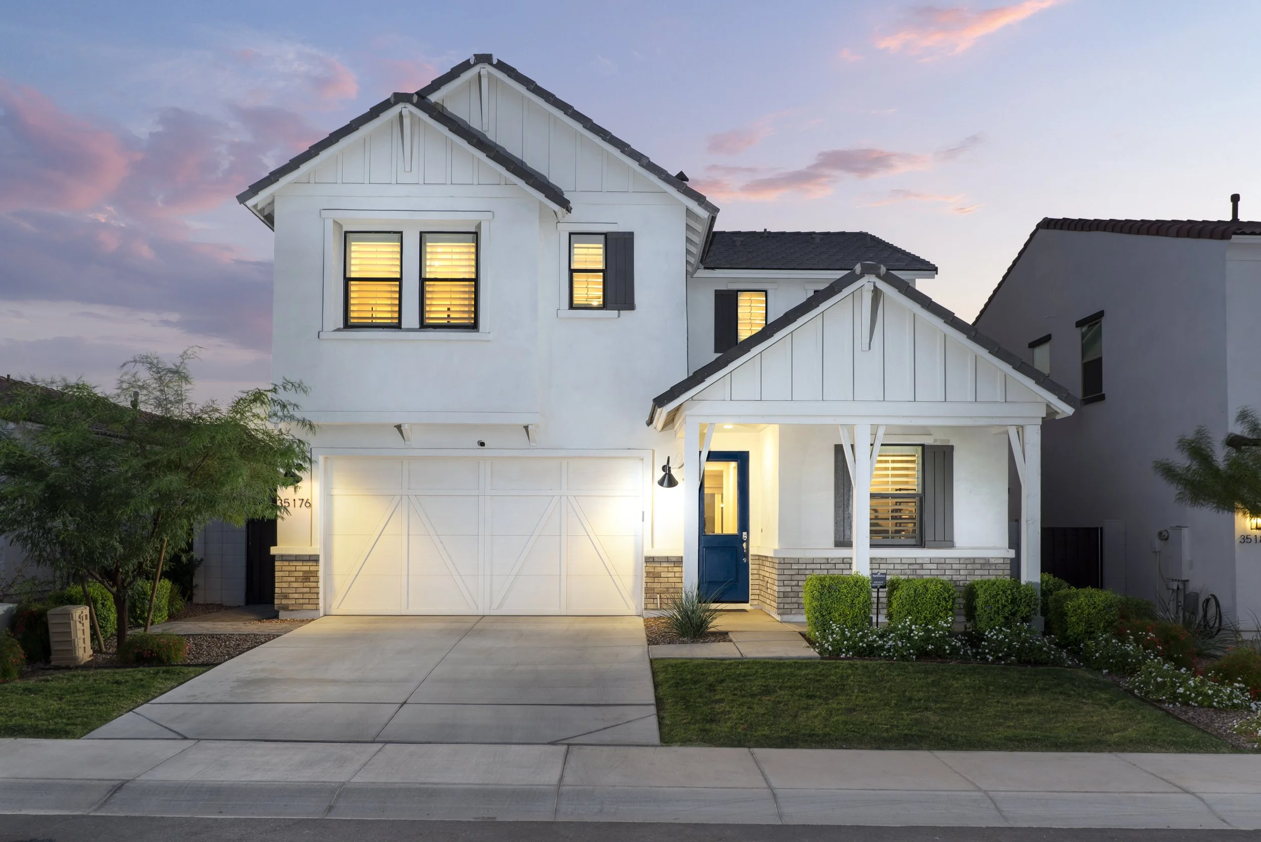Modern two-story white house with a blue front door, illuminated windows, and a small front porch with a gabled roof, surrounded by a well-kept lawn and greenery at sunset.