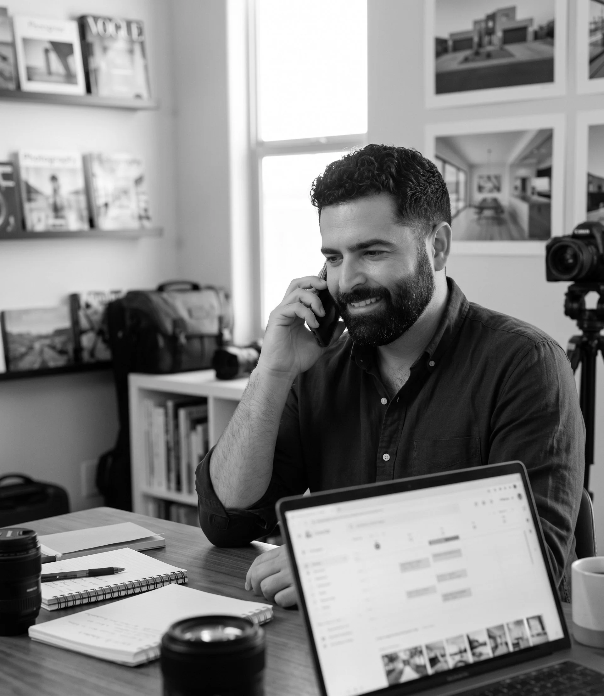 Sean Colon talking on a mobile phone while sitting at a desk in a room with artwork on the wall and a window. The desk has notebooks, a pen, a camera lens, and a coffee cup. A laptop screen displays an email inbox in Phoenix Arizona.