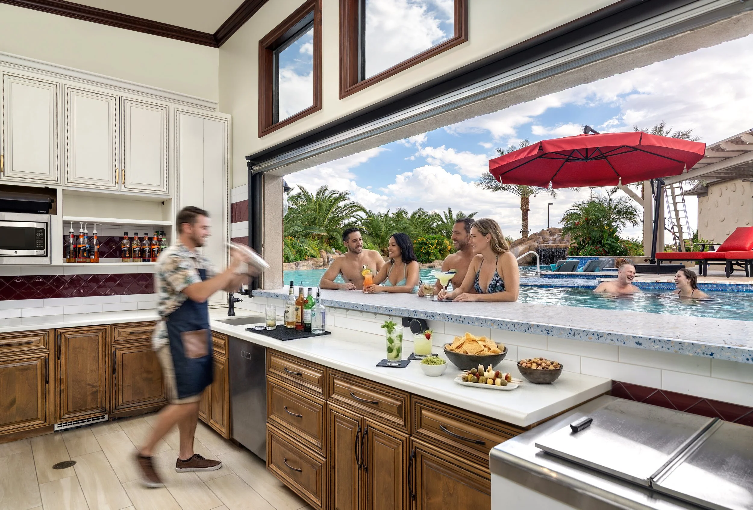 People enjoying a poolside gathering with drinks and snacks while a bartender prepares beverages in an outdoor covered bar area with large windows and a view of the pool and palm trees.