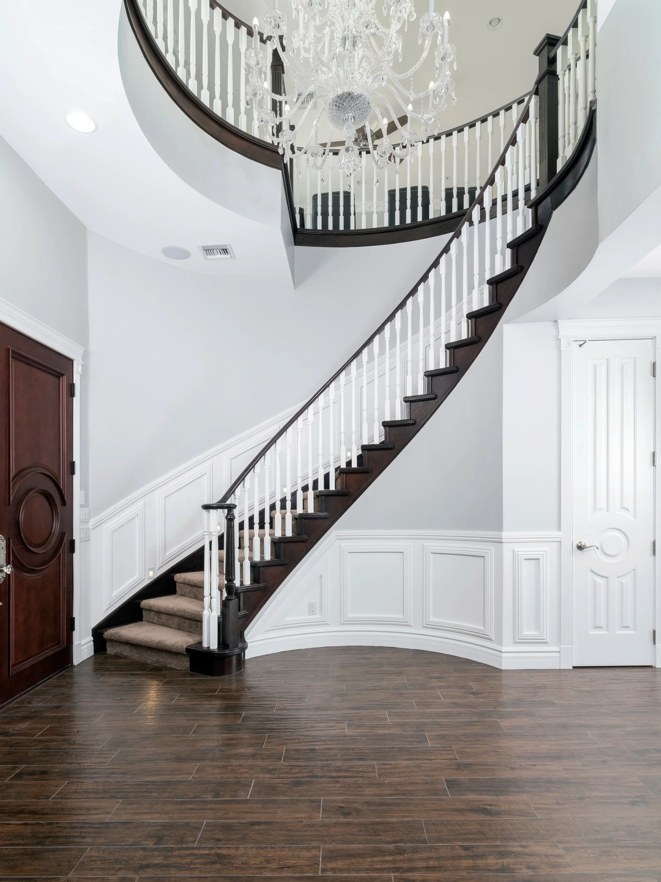 A staircase with dark wooden steps and white balusters curves upward to a second floor, where a crystal chandelier hangs from the ceiling. The lower floor has dark wood flooring, white wainscoting, and two white doors, one on each side of the staircase.