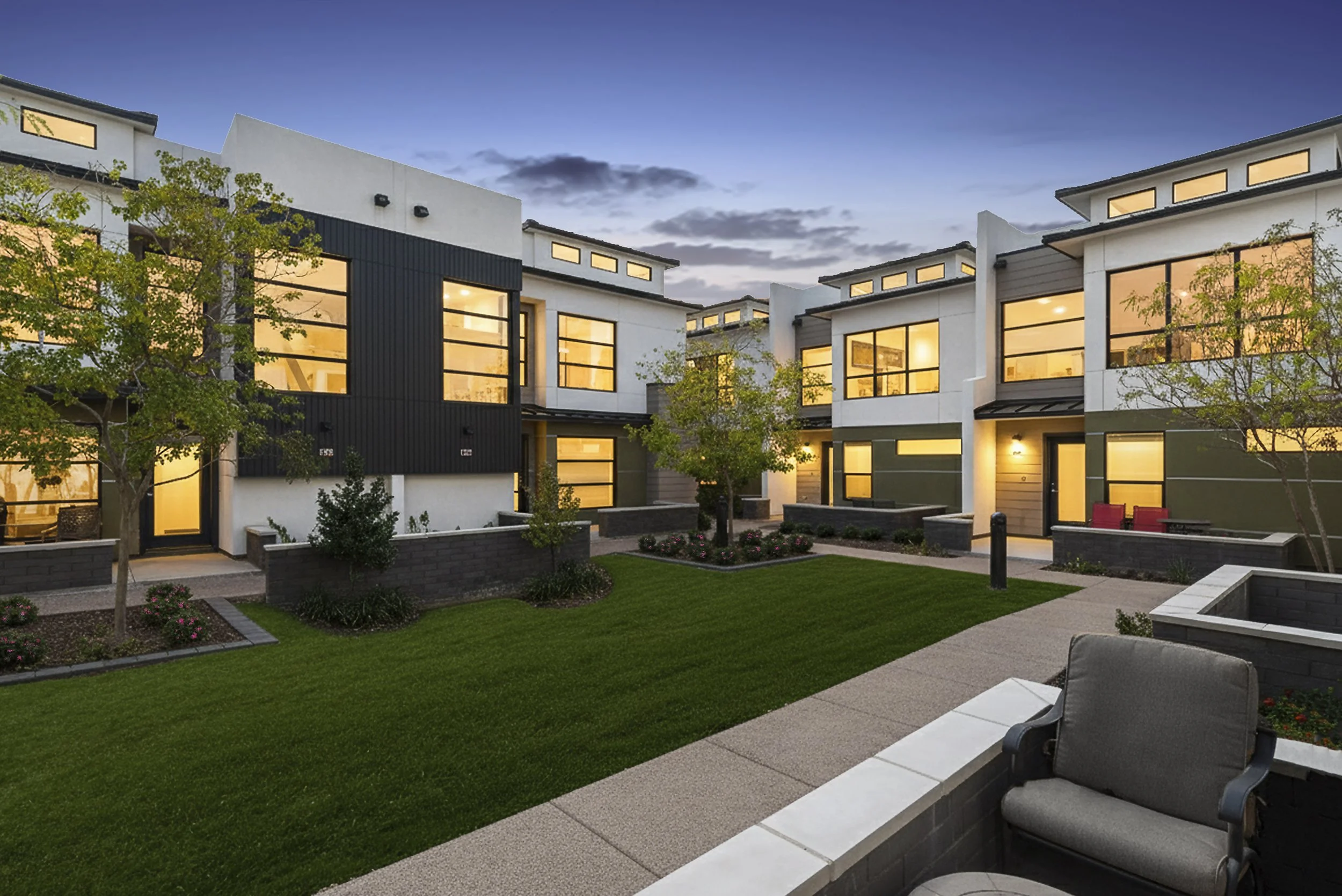 Modern townhouses with large windows and front yards, illuminated at dusk, with trees, grass, and a patio area with chairs.