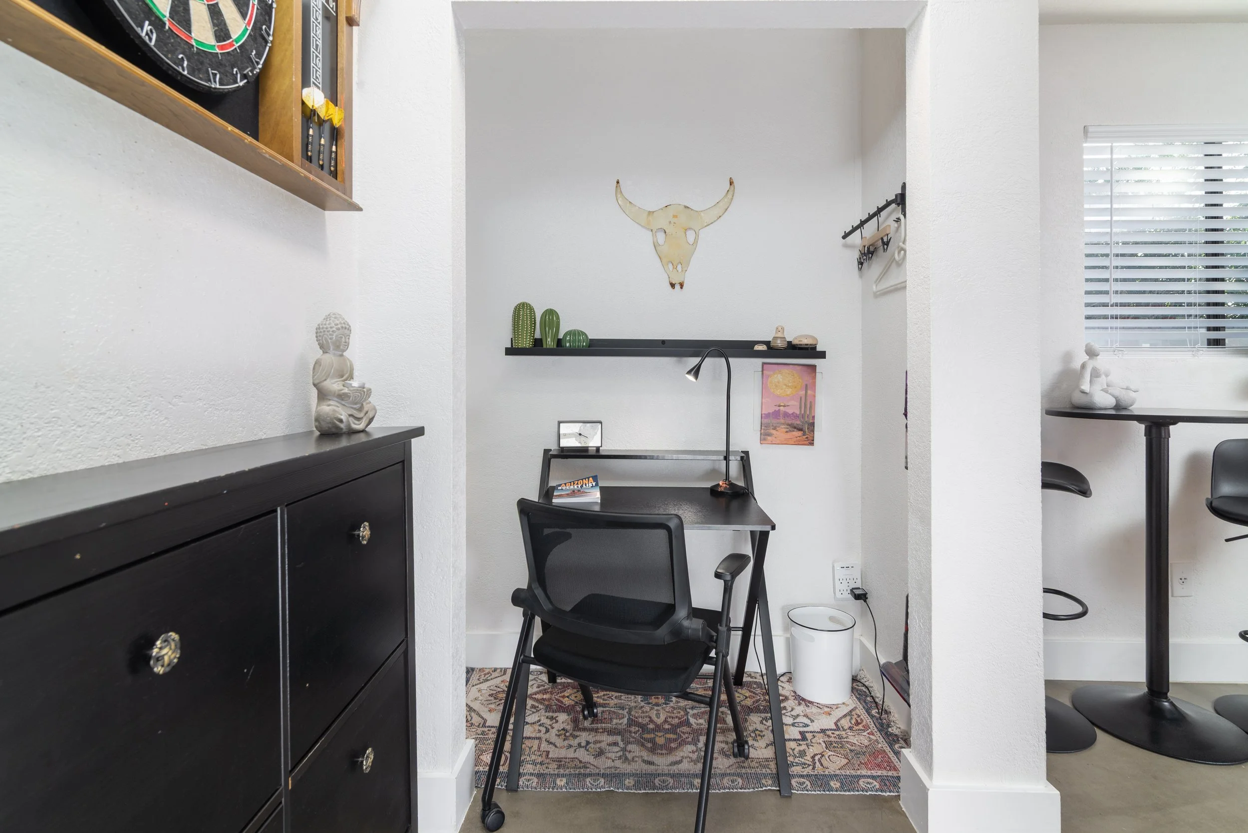 A small office space with white textured walls, black furniture, and decorative items. The back wall features a bull skull with horns, above black shelves with small cactus plants and a framed desert landscape art. To the left, a black dresser with decorative Buddha statue and a dartboard is visible. To the right, part of a window with blinds and a round high table with a sculpture are seen. A black office chair faces a small desk with a desk lamp, and there is a trash can below it.