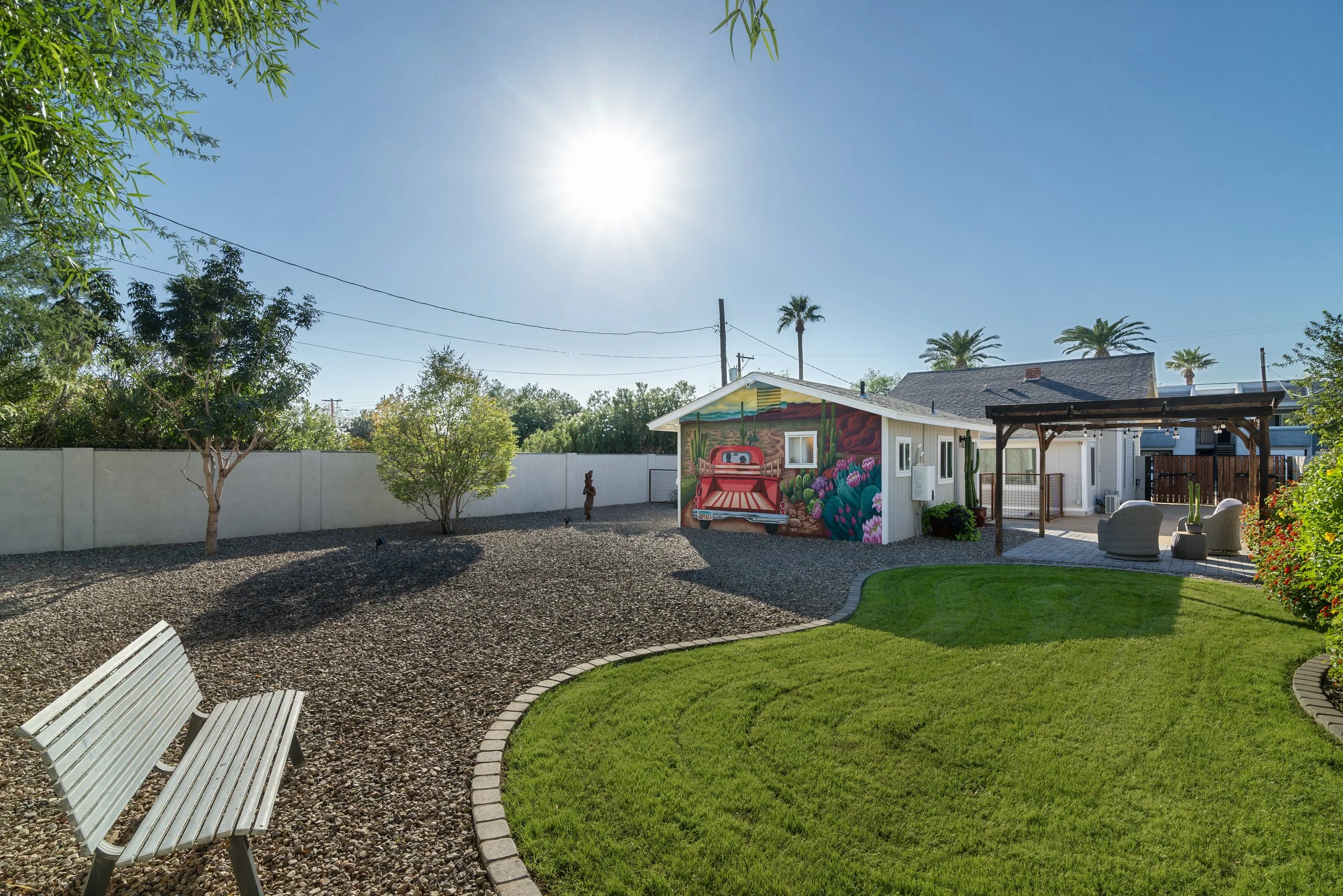 A backyard with a well-maintained lawn, a painted shed featuring a cactus and desert scene, white benches, a patio area with outdoor furniture, tall palm trees, and a white fence under a clear sunny sky.