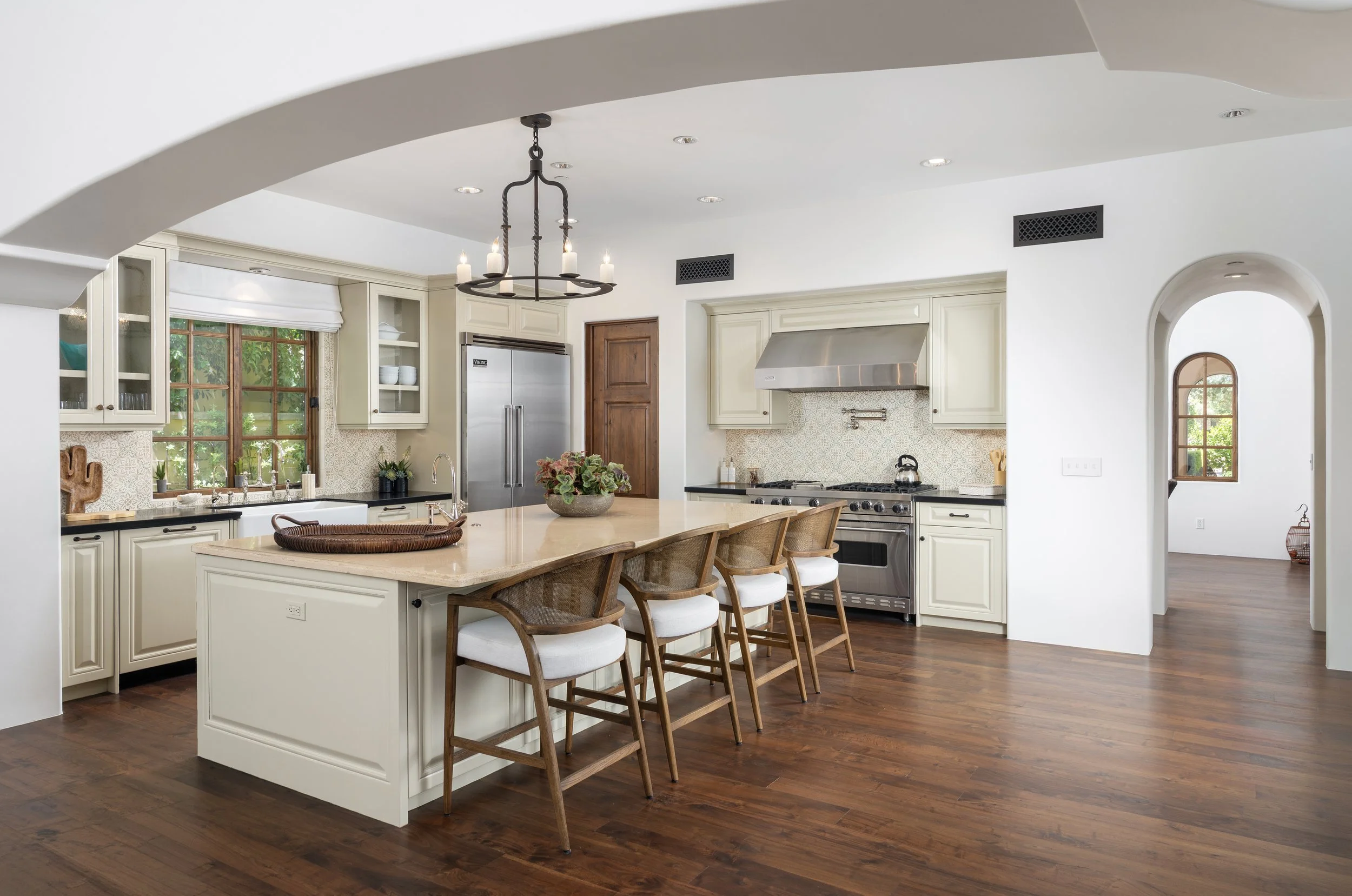 Bright, modern kitchen with white cabinets, wooden accents, a large kitchen island with seating, stainless steel appliances, a chandelier, and a wooden floor.