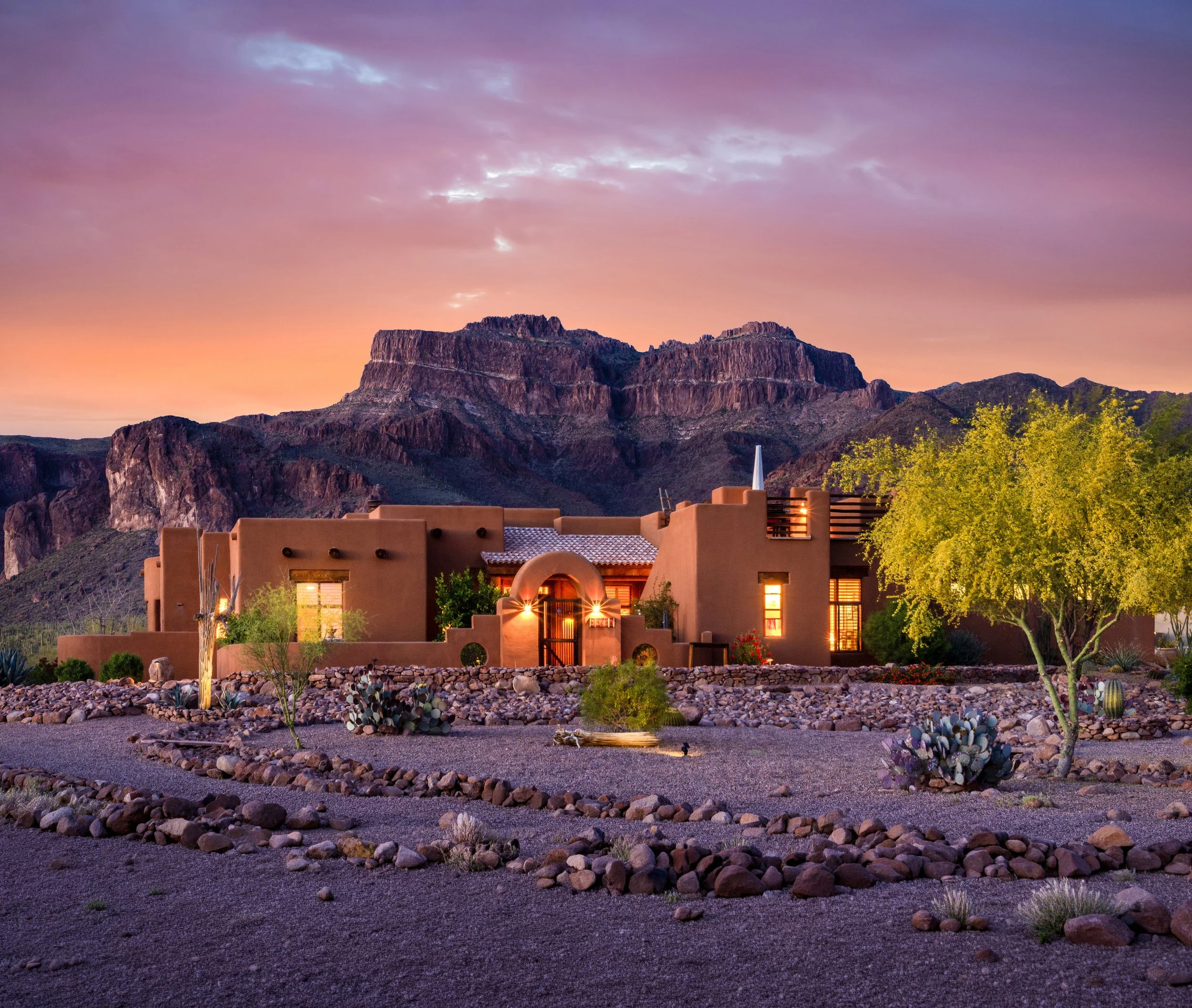 Desert landscape with the Superstition mountains under a colorful sunset sky, foreground features a landscaped yard with rocks, plants, and a desert-style adobe home in Superstition Highlands neighborhood in Apache Junction, Arizona.