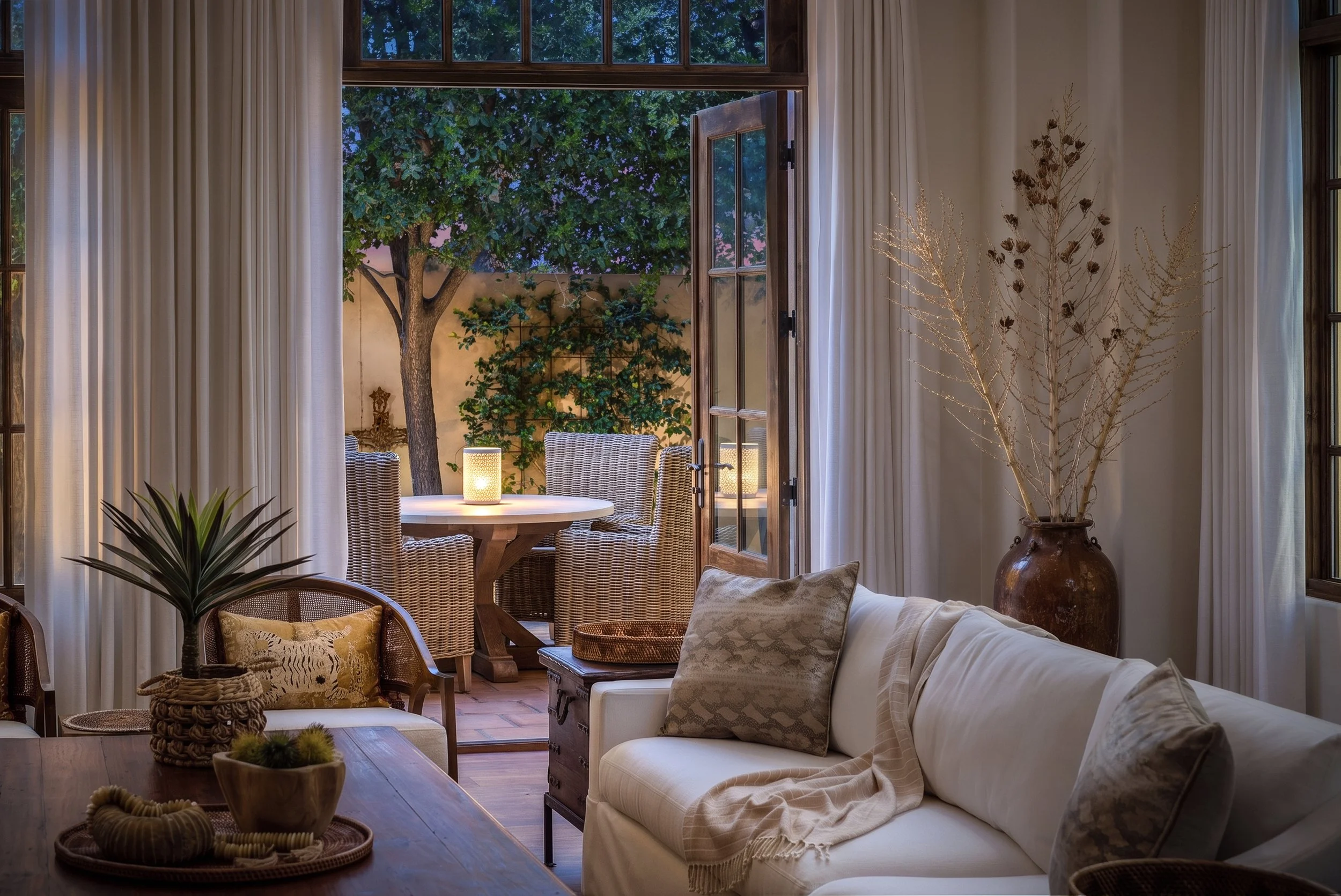 Interior view of a living room with an open door leading to a patio with outdoor furniture and a large tree. The room has cream-colored curtains, decorative plants, and cushions on a sofa.