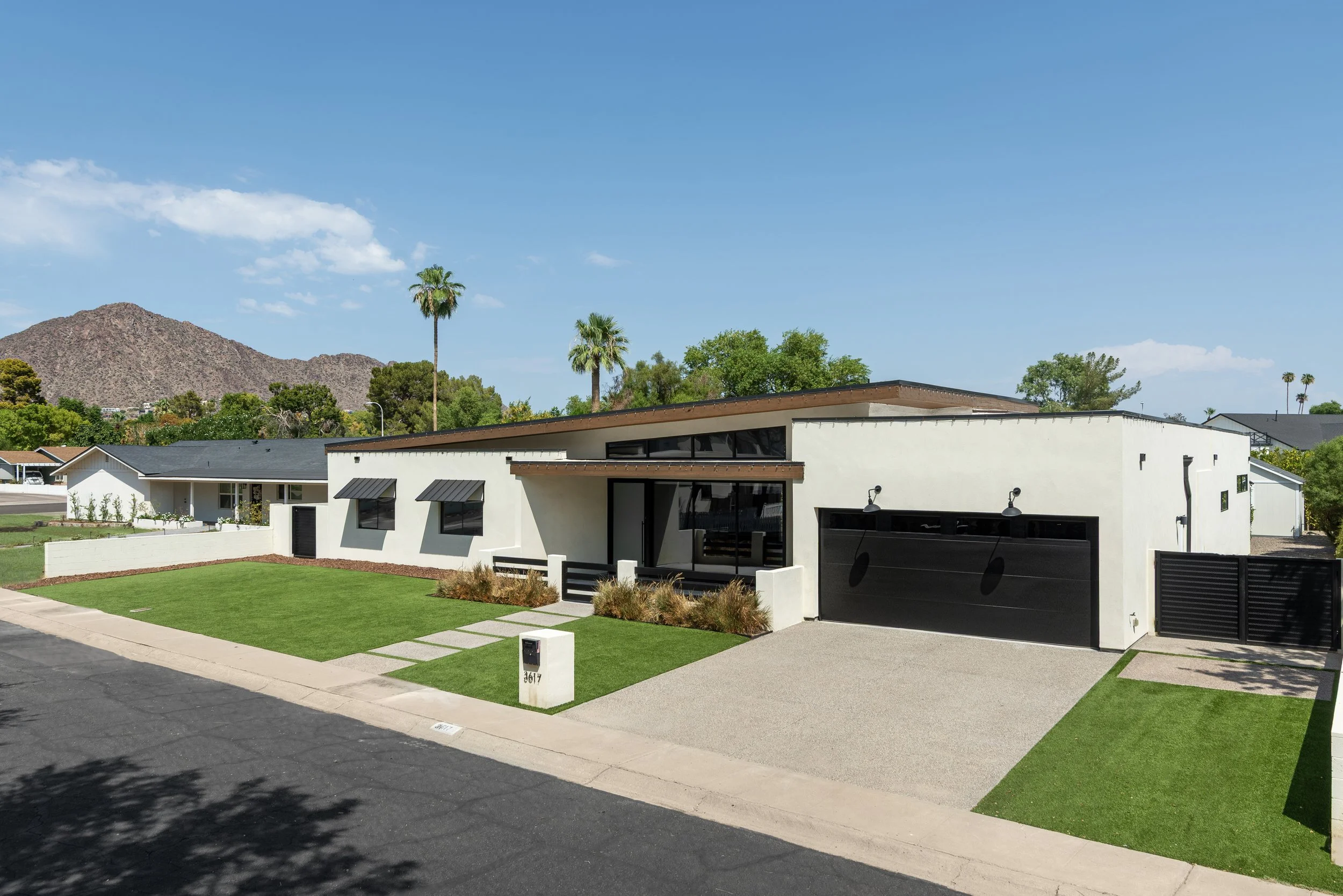 Modern single-story house with white exterior, black garage door, and small front yard with green grass and pathway, mountain and palm trees in background, sunny sky in arcadia, phoenix, arizona.