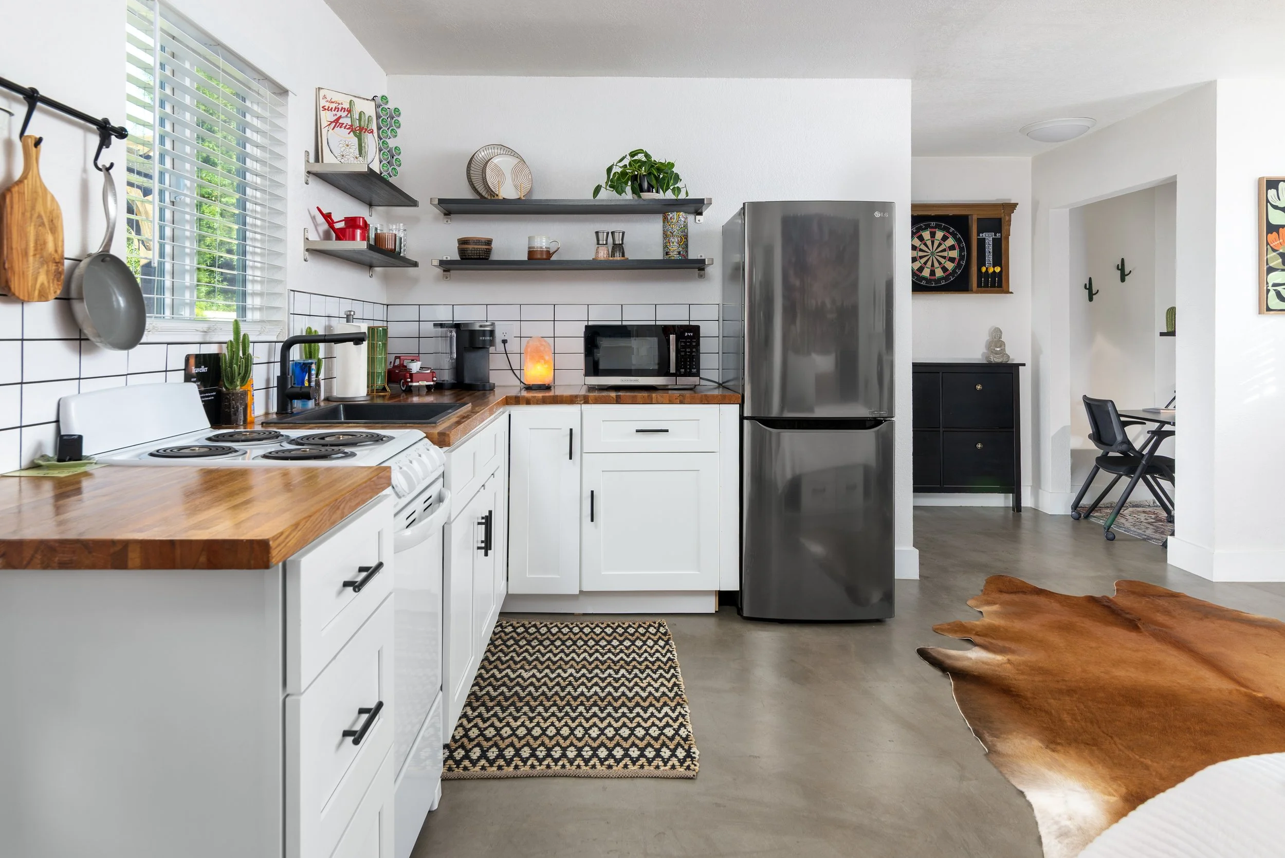 A modern kitchen with white cabinets, open shelves, stainless steel refrigerator, microwave, and a coffee maker. There are decorative plants, a salt lamp, and wall art featuring cacti. A black and cream woven rug on the floor and a wooden cowhide rug near the dining area.