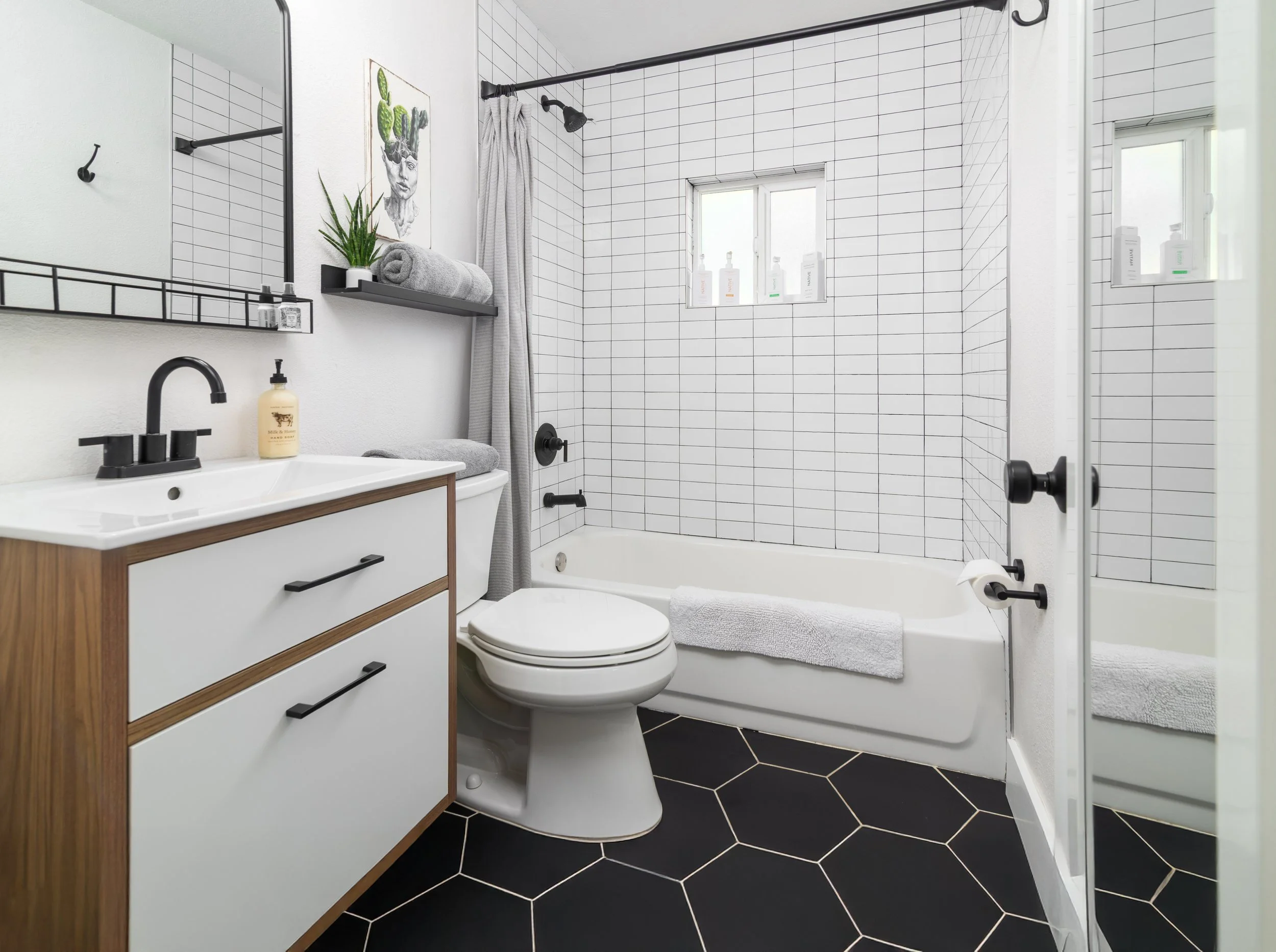 A modern bathroom with white tiled shower area, black fixtures, a vanity with a black faucet, a toilet, black hexagonal floor tiles, a mirror, a shelf with towels and plants, and a window with styling bottles.