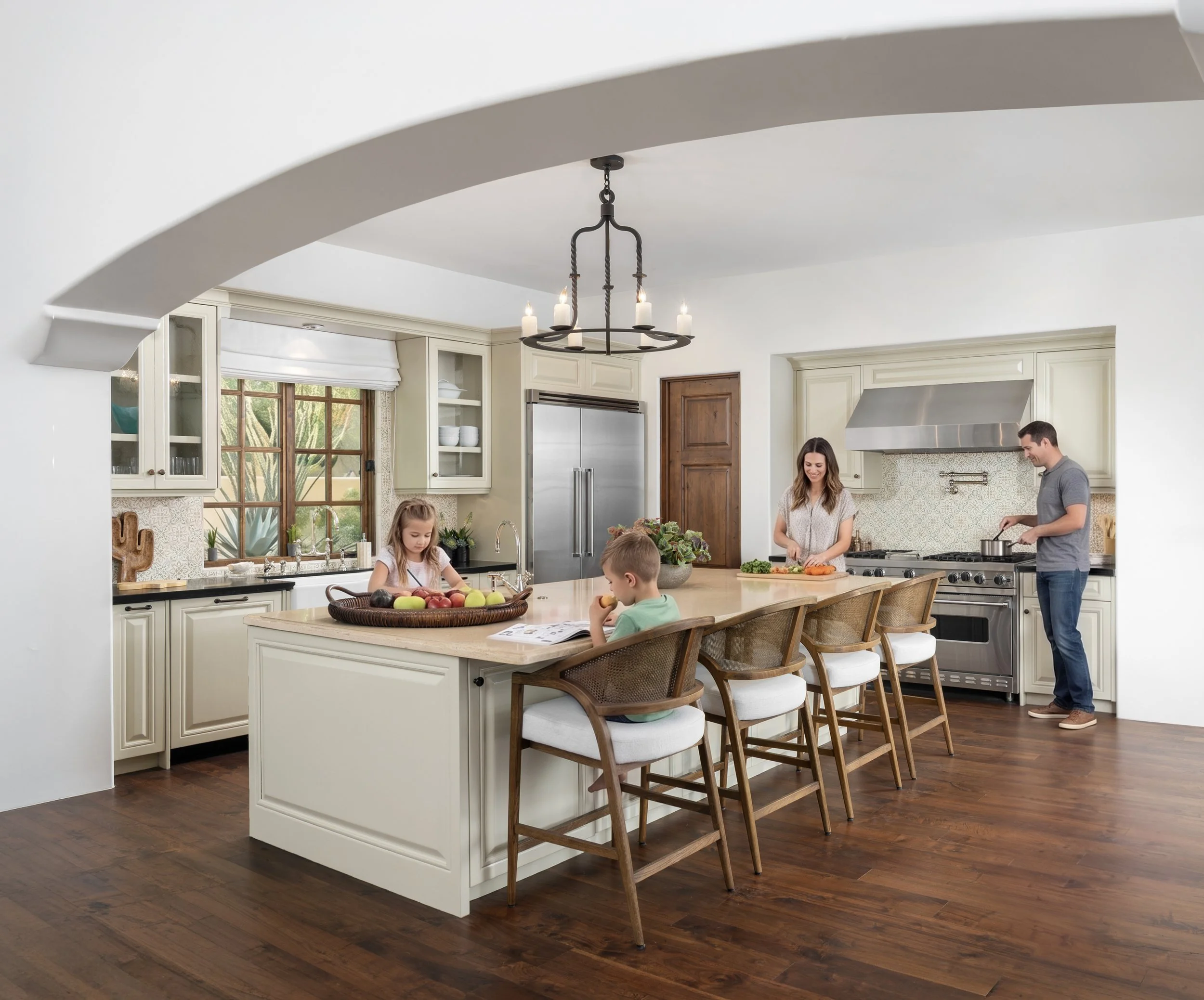 A spacious kitchen with white cabinets, a large kitchen island with a light-colored countertop, and wood flooring. There are three children seated at the island and two adults standing and preparing food. A large window with a white Roman shade allows natural light to illuminate the space. A modern black chandelier hangs above the island.