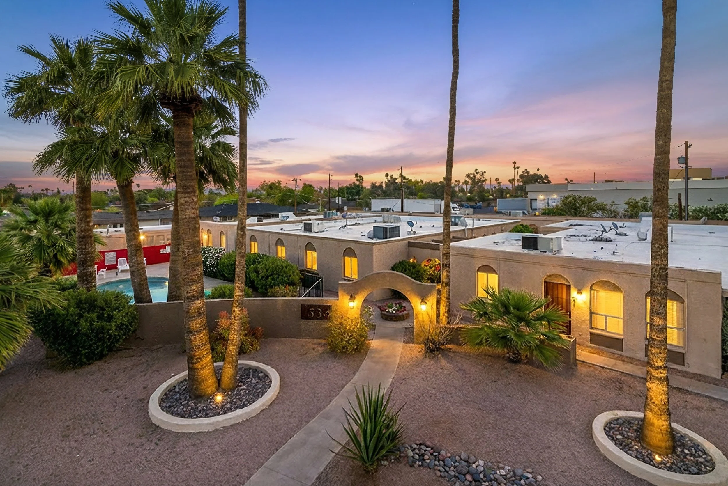A view of a suburban neighborhood at dusk with a sunset sky, tall palm trees in the foreground, and a beige apartment complex with arched windows and rooftop air conditioning units. There is a small landscaped area with a circular flower bed, bushes, and a patio with chairs near a swimming pool.