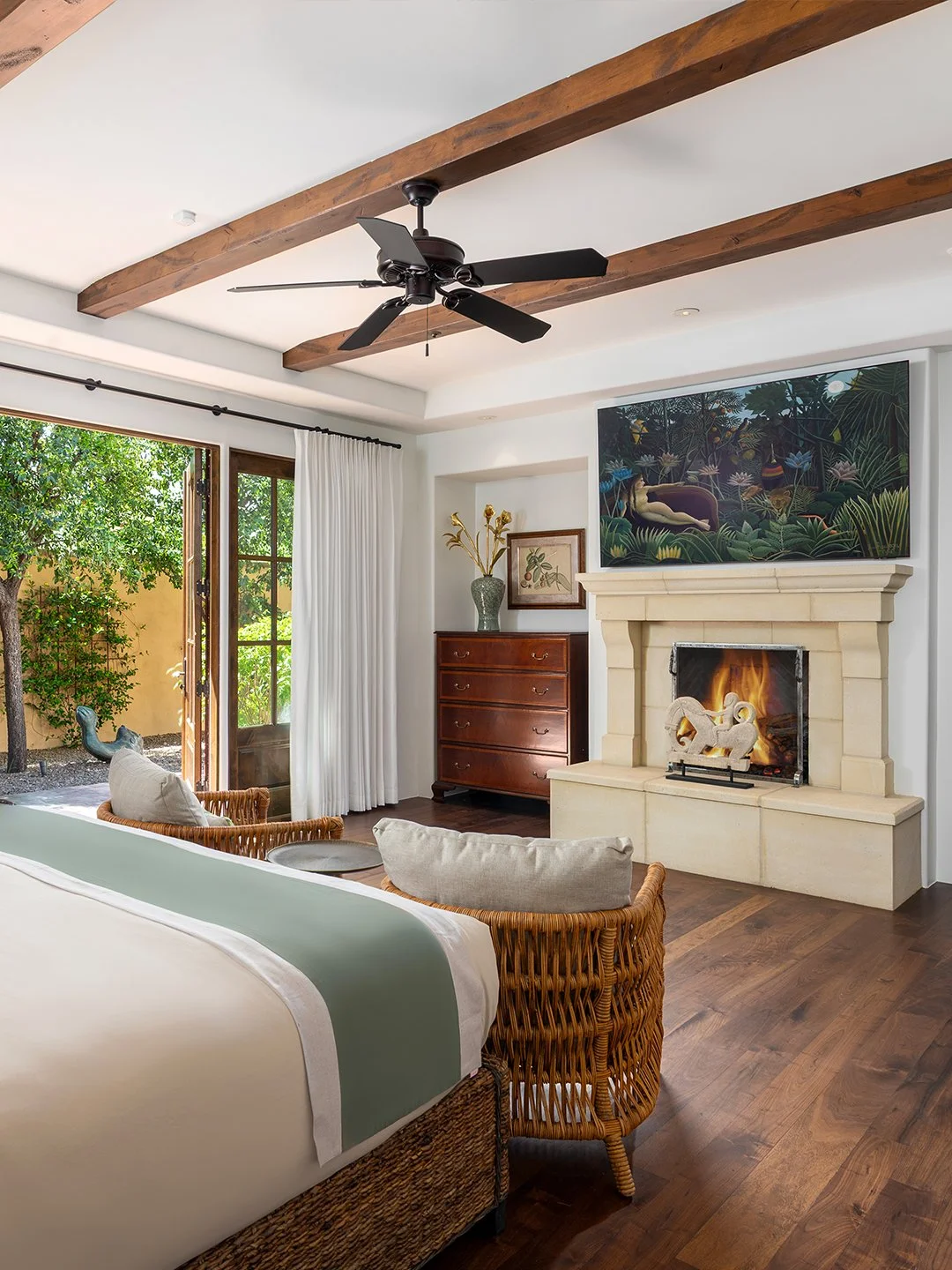Living room with a fireplace, wooden ceiling beams, a ceiling fan, large window with white curtains, wicker armchairs, a bed with a green stripe, a wooden dresser, and a framed painting above the fireplace.