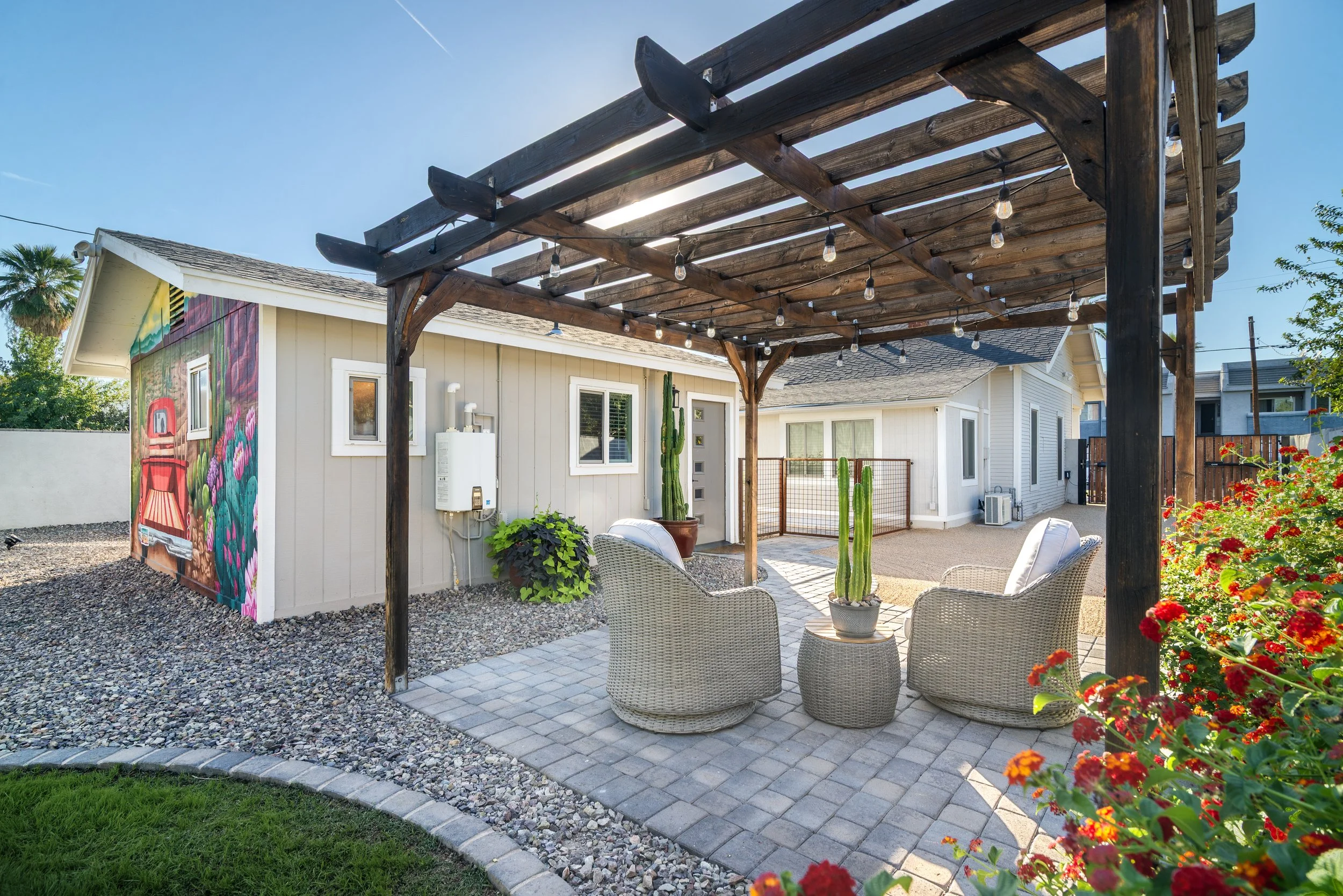 A backyard patio with a wooden pergola, string lights, outdoor furniture, potted plants, colorful flowers, and a mural on a side building, under a clear blue sky.