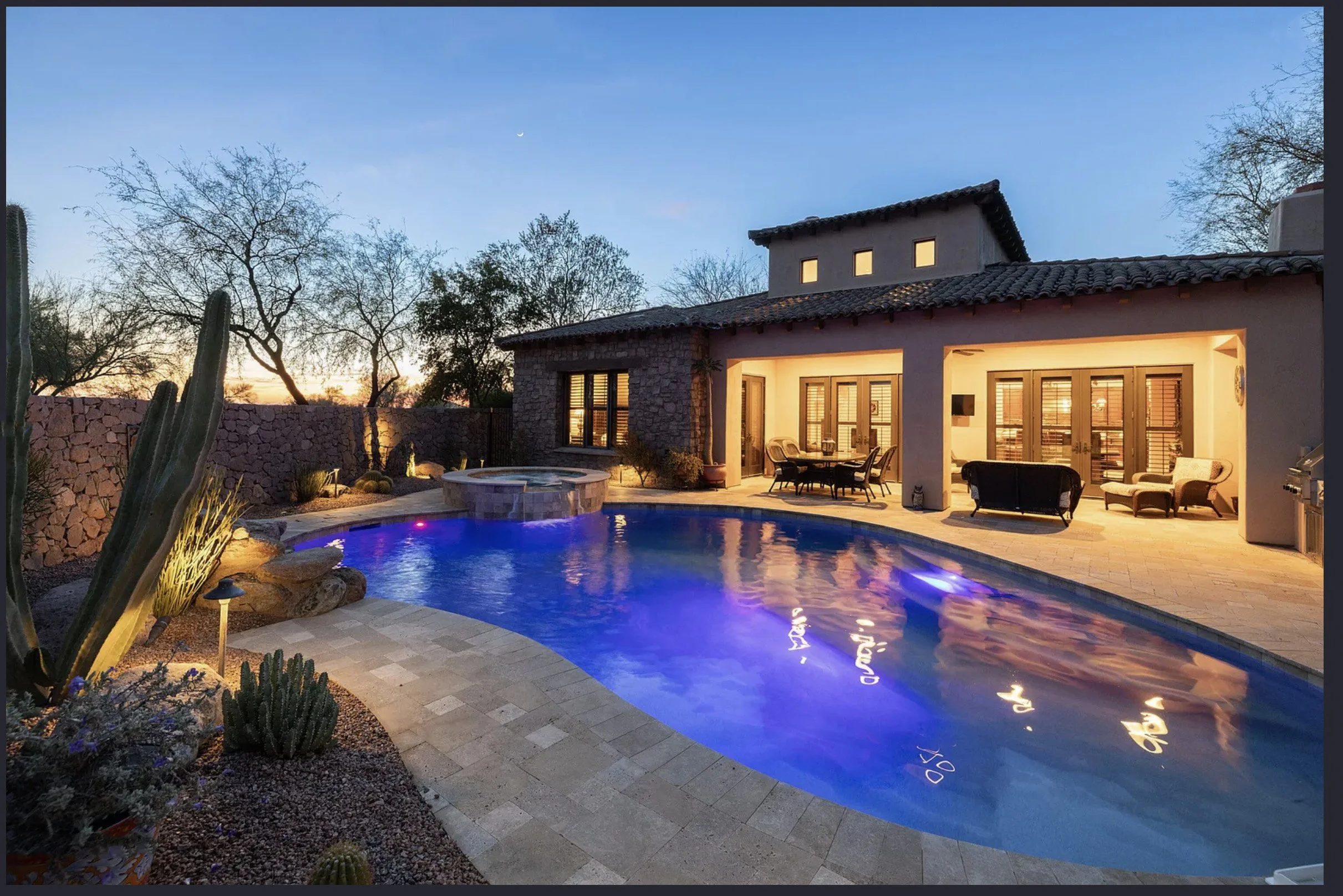 A backyard with a swimming pool with underwater lights, outdoor seating area inside a covered patio, desert plants, cacti, and a stone wall fence, during twilight evening .