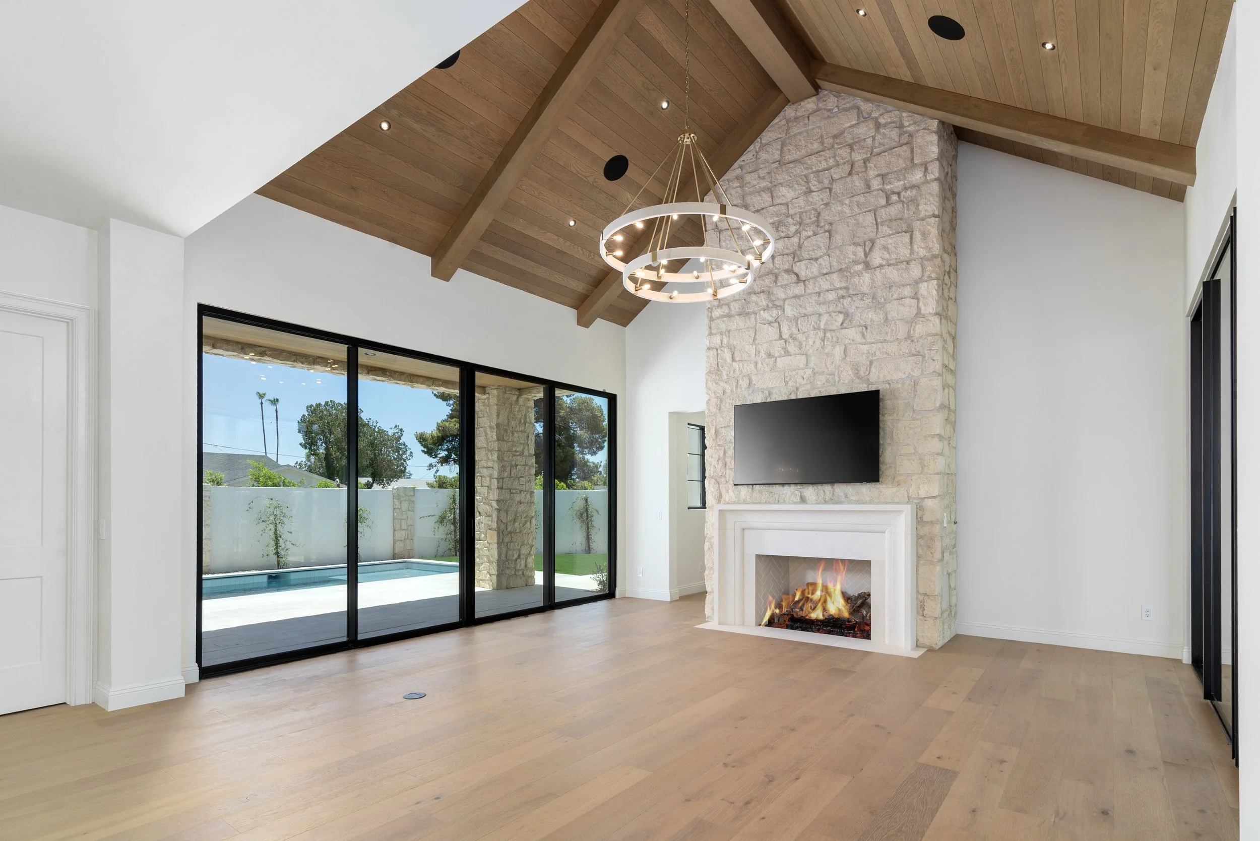Modern living room with a stone fireplace, large sliding glass doors leading outside, and a high wooden vaulted ceiling with a chandelier.