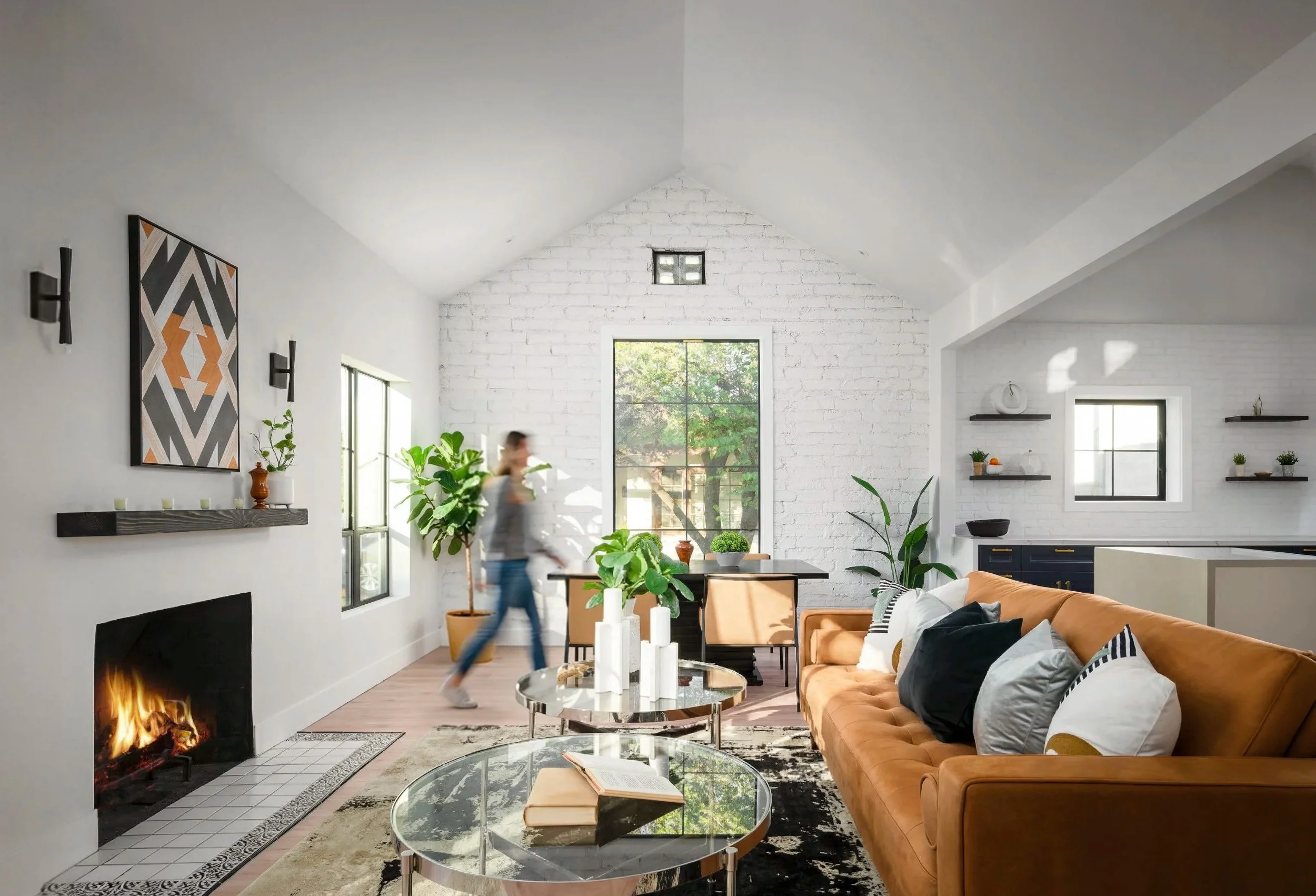 Bright living room with white brick walls, large windows, and a high ceiling. Features a leather sofa with cushions, a fireplace, and potted plants. A person is walking in the background near a dining table.