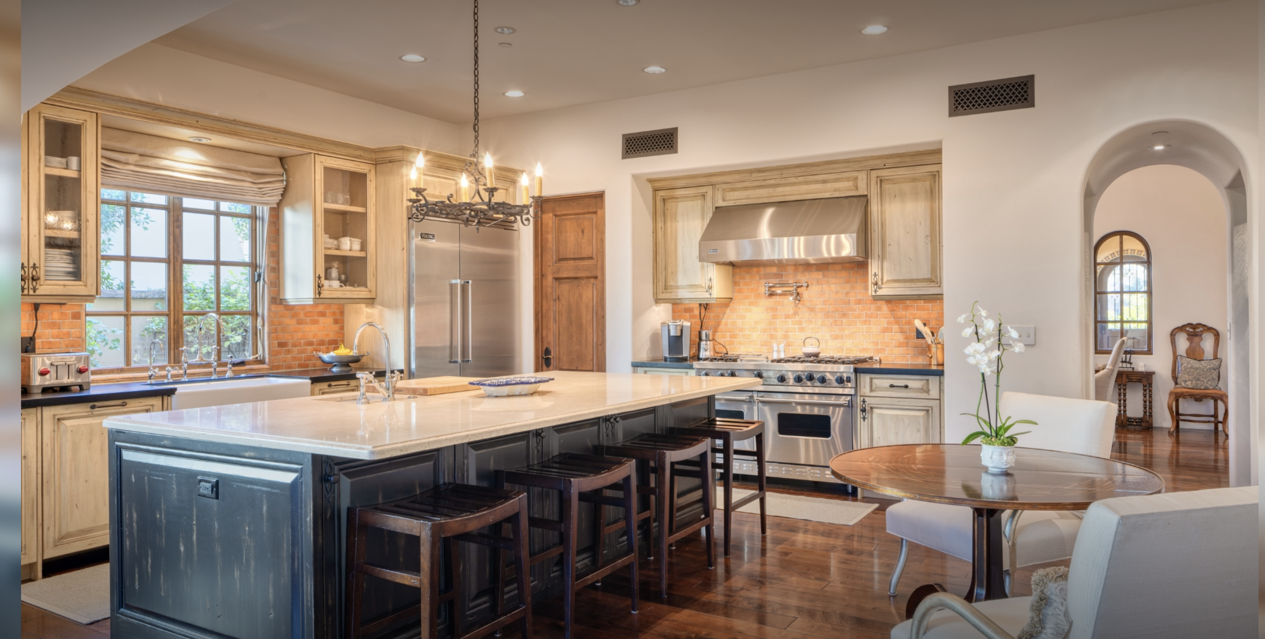 A spacious kitchen with a large island, wooden cabinetry, stainless steel appliances, a chandelier, and a dining area with a white sofa and round table with a flower arrangement.