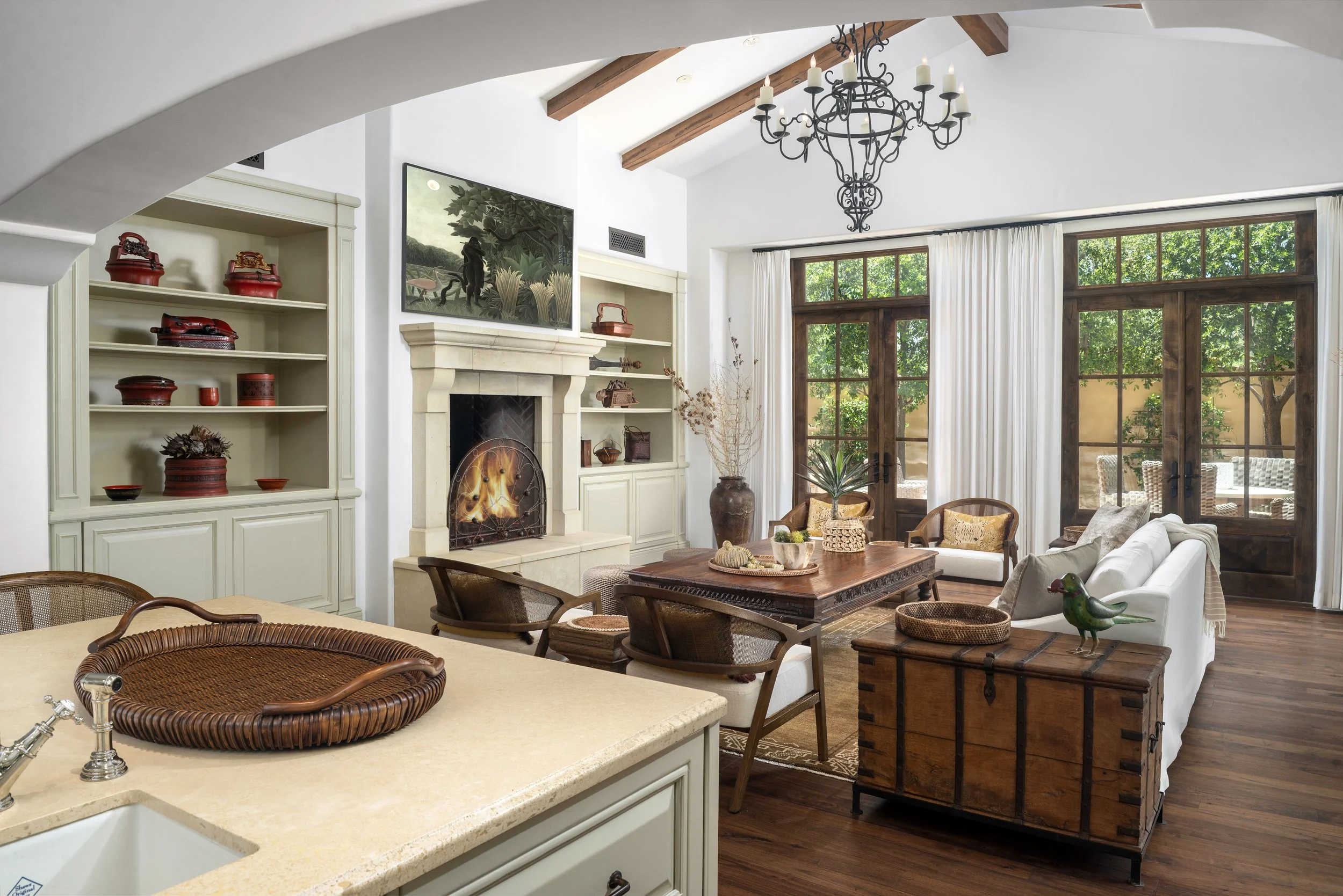 Bright living room with white walls, exposed wooden beams, a chandelier, a fireplace, built-in white shelves with decorations, large windows with white curtains, and outdoor view of trees, with furniture including a white sofa, wooden chairs, and a wooden chest.