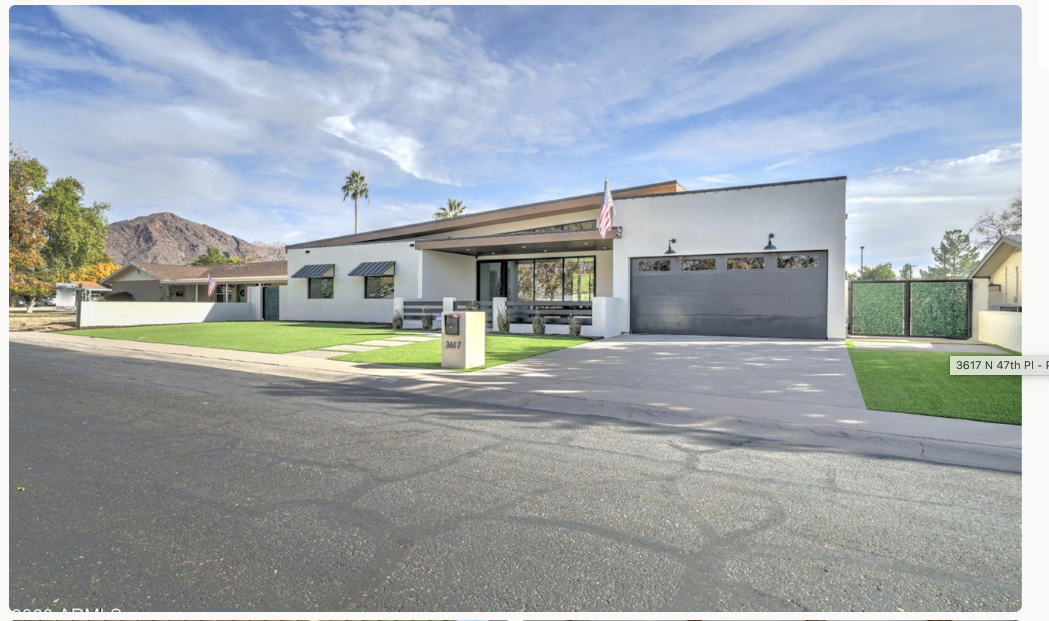A modern single-story house with a white exterior, black garage door, and a front yard with green artificial grass and concrete pathway. An American flag hangs from the front of the house beneath a small roof. The house is situated on a street with mountainous terrain in the background and a clear blue sky.