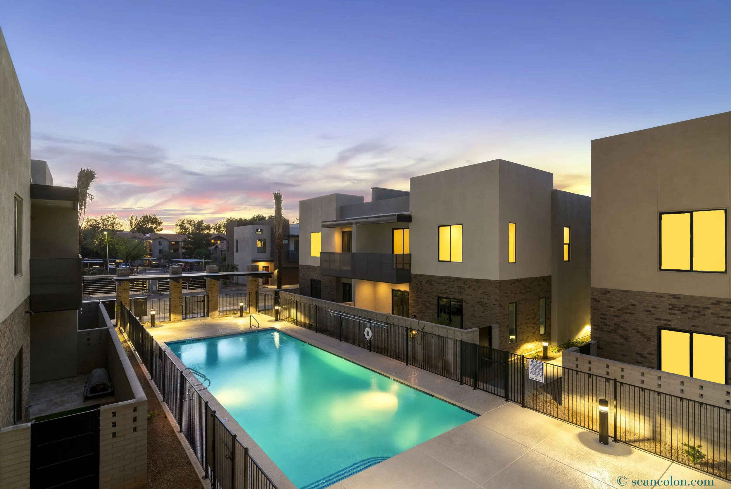 Apartment complex with a swimming pool, lit by outdoor lighting at sunset, with modern buildings and a colorful sky in the background.