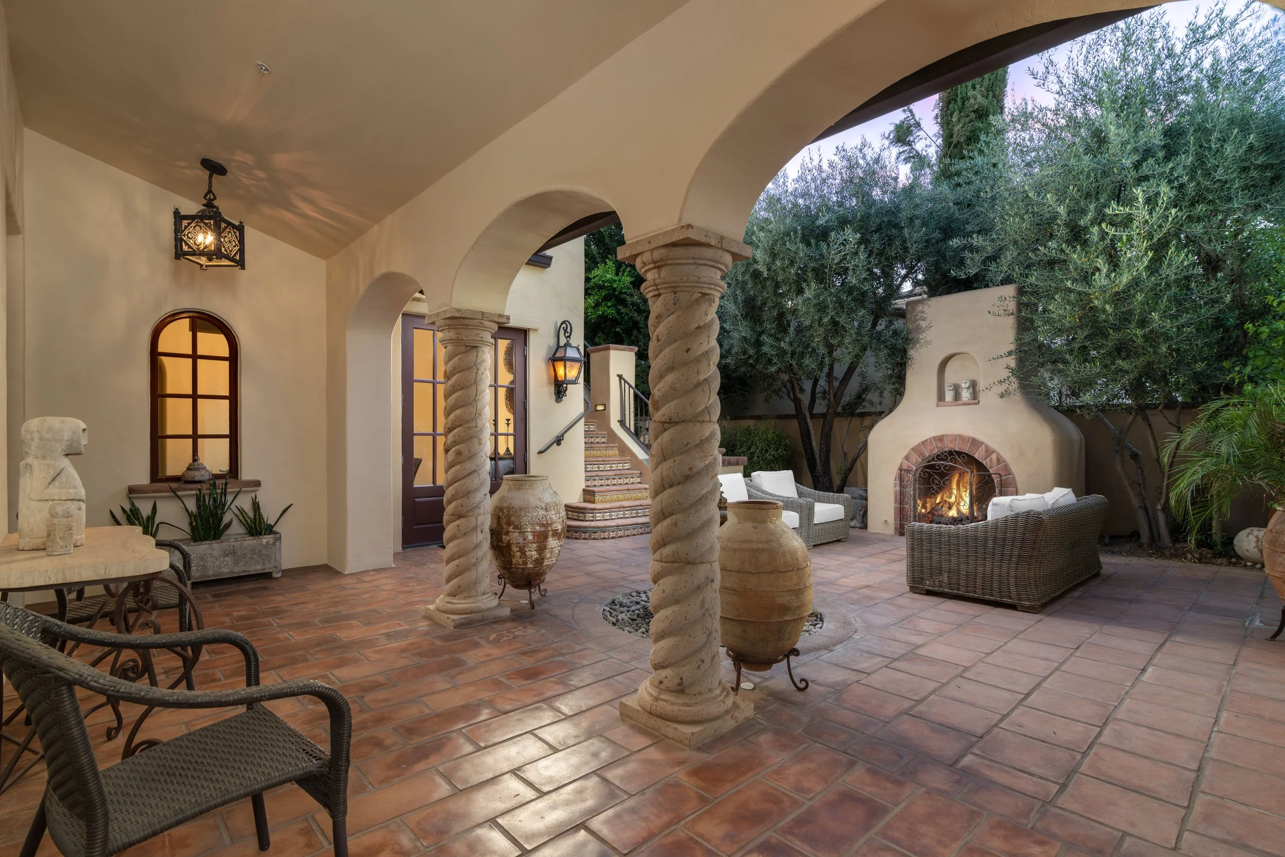 Patio area with terracotta tile flooring, arched ceiling, and stone columns. There are wicker chairs, a fireplace with a fire burning, and greenery including trees.