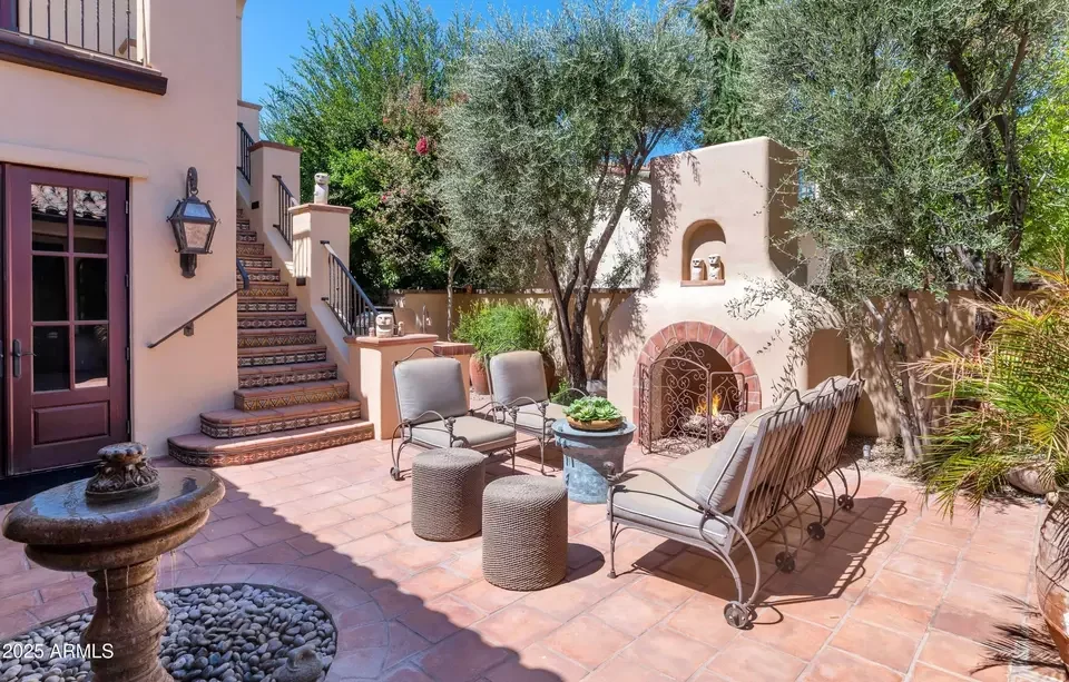 Outdoor patio with outdoor seating, wall fireplace, potted plants, and trees, on a sunny day.
