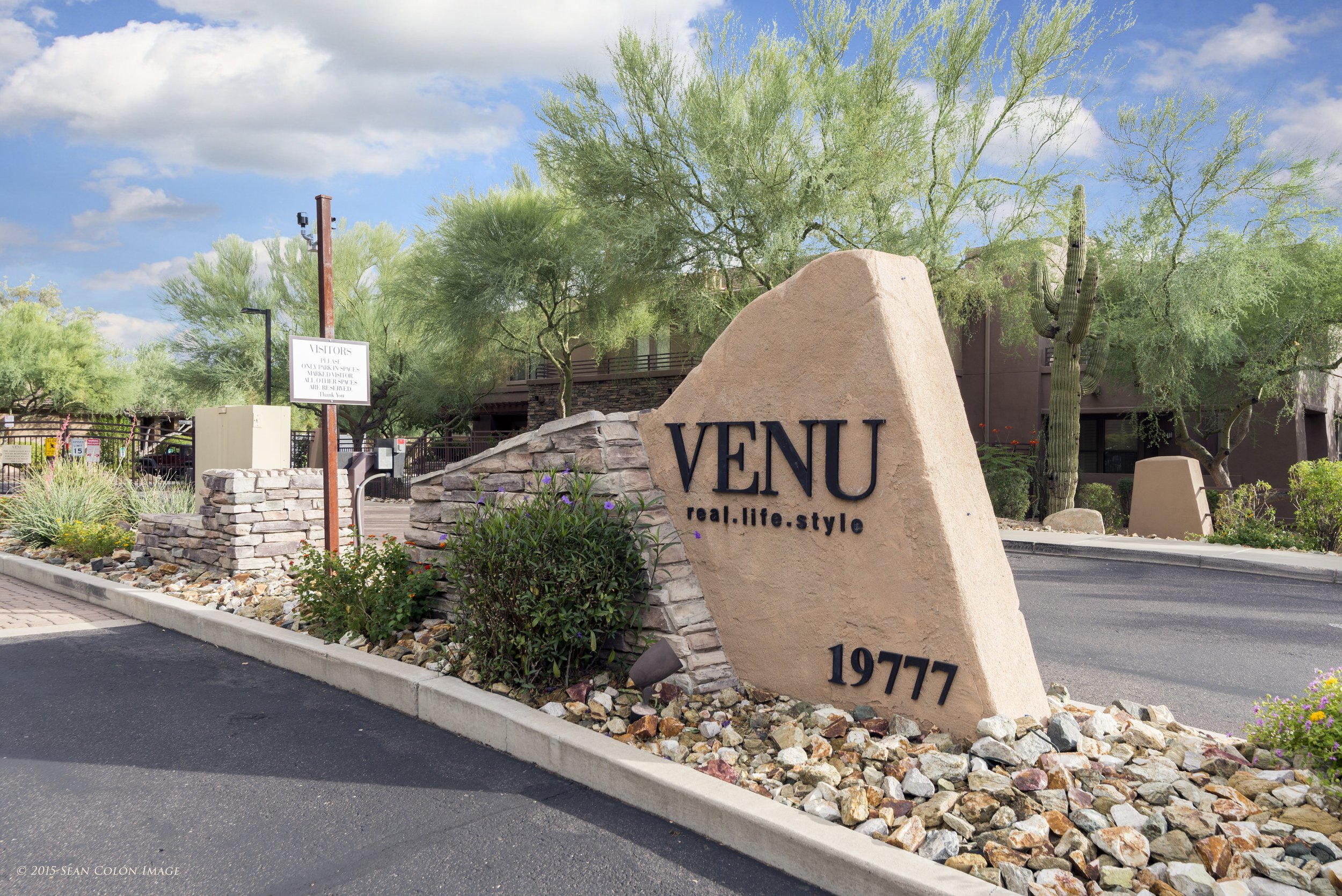 A large stone sign reading VENU with the tagline 'real.life.style' and the number 19777. The sign is surrounded by desert plants and rocks, with a background of desert trees, cacti, and a partly cloudy sky.