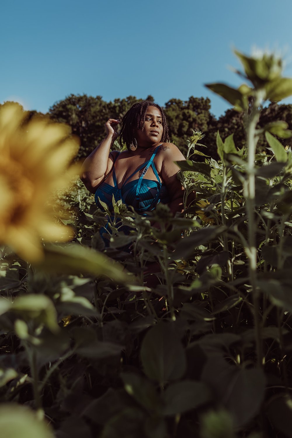  Black woman in lingerie is covered by sunflower stems in an outdoor photoshoot while standing among the stalks 