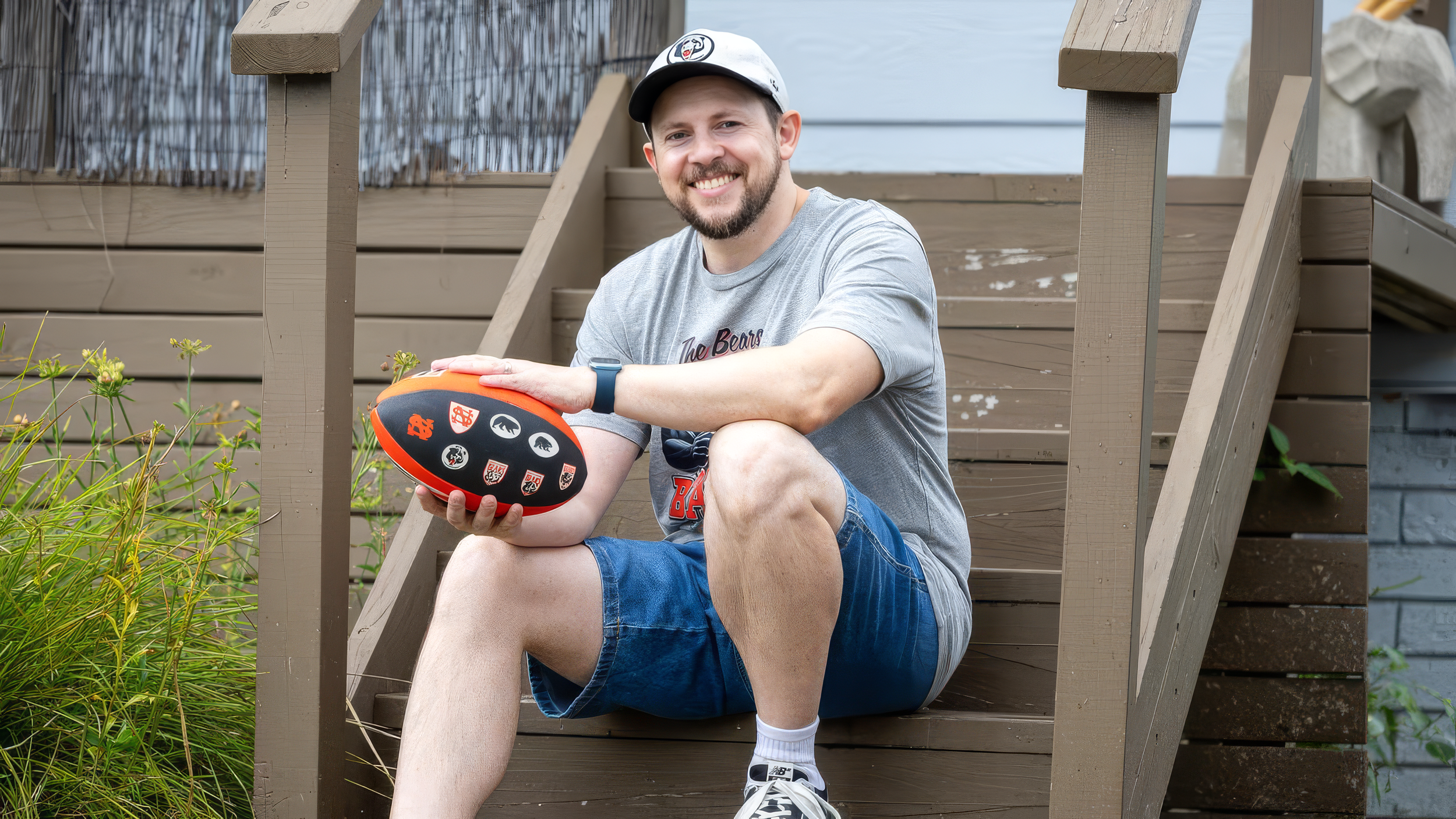 A smiling man sitting on wooden stairs outdoors, holding a football with team logos. He wears a gray t-shirt, blue shorts, white socks, sneakers, a cap, and a smartwatch.