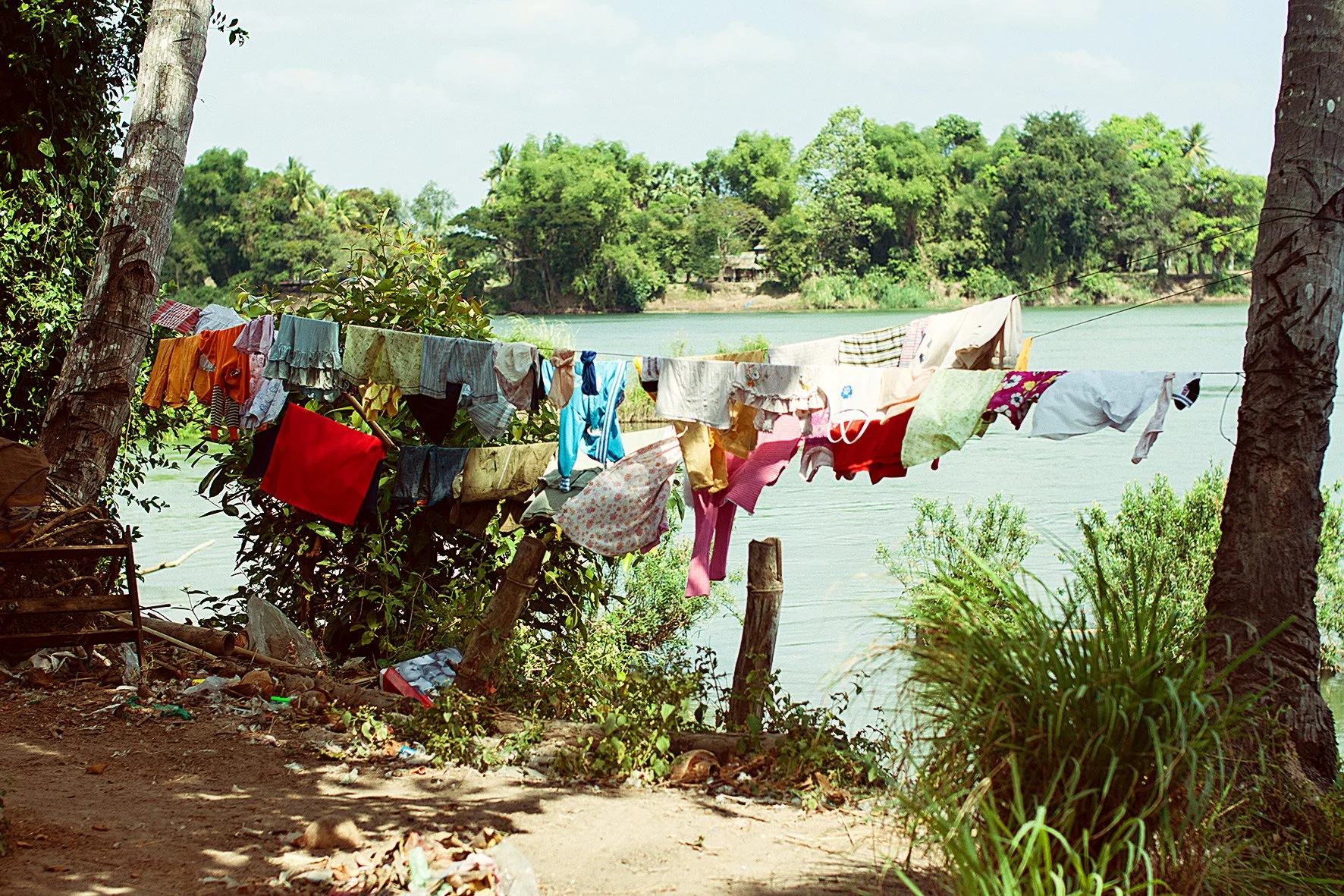 Linge étendu sur une corde à linge entre deux arbres près d'une rivière, avec une végétation luxuriante en arrière-plan.
