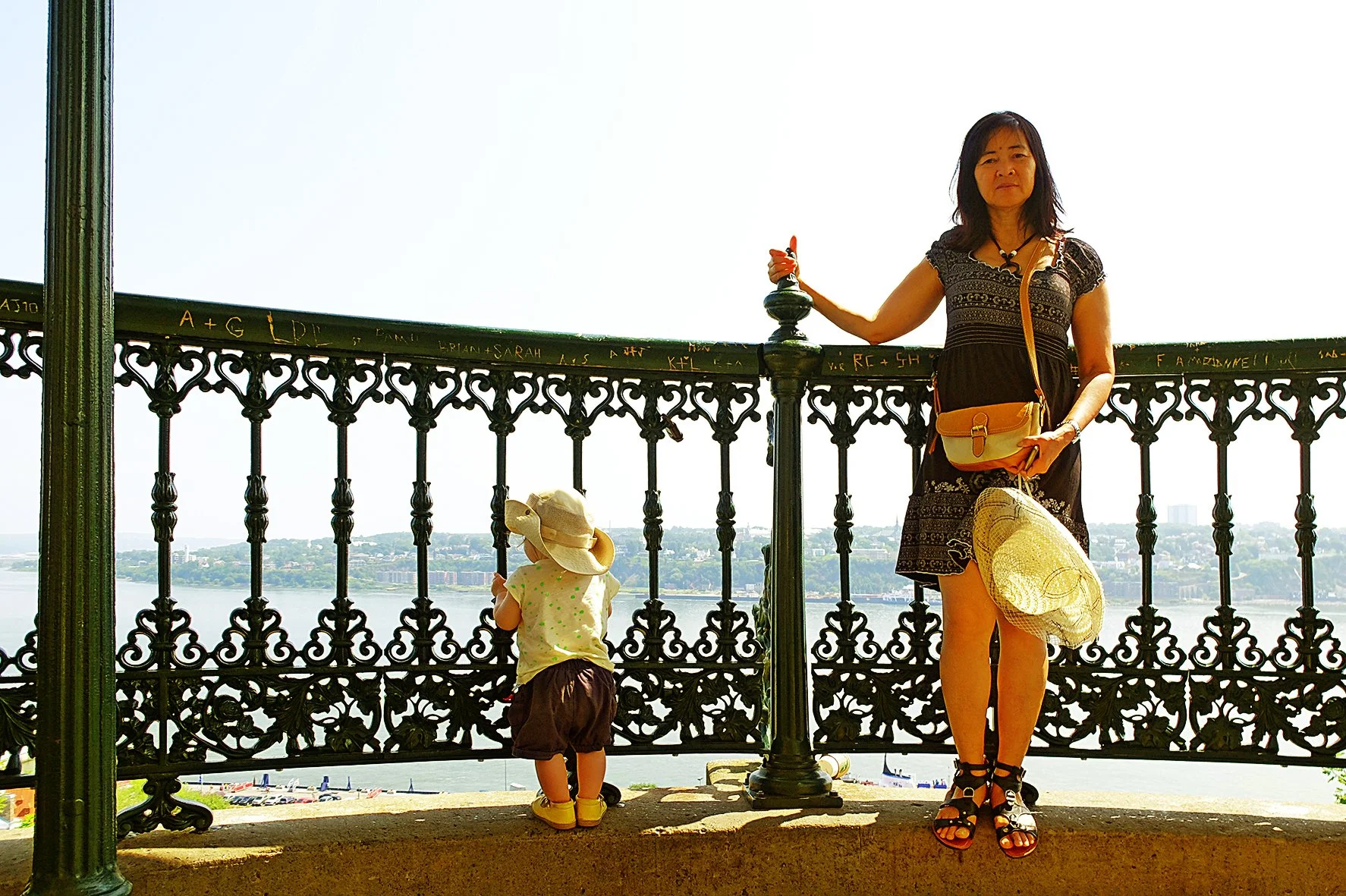 Une femme et un enfant regardant un panorama depuis une balustrade en fer forgé dans une zone urbaine avec un lac en arrière-plan. La femme porte une robe noire, des sandales noires, un sac à bandoulière beige, et tient un chapeau de paille. L'enfant