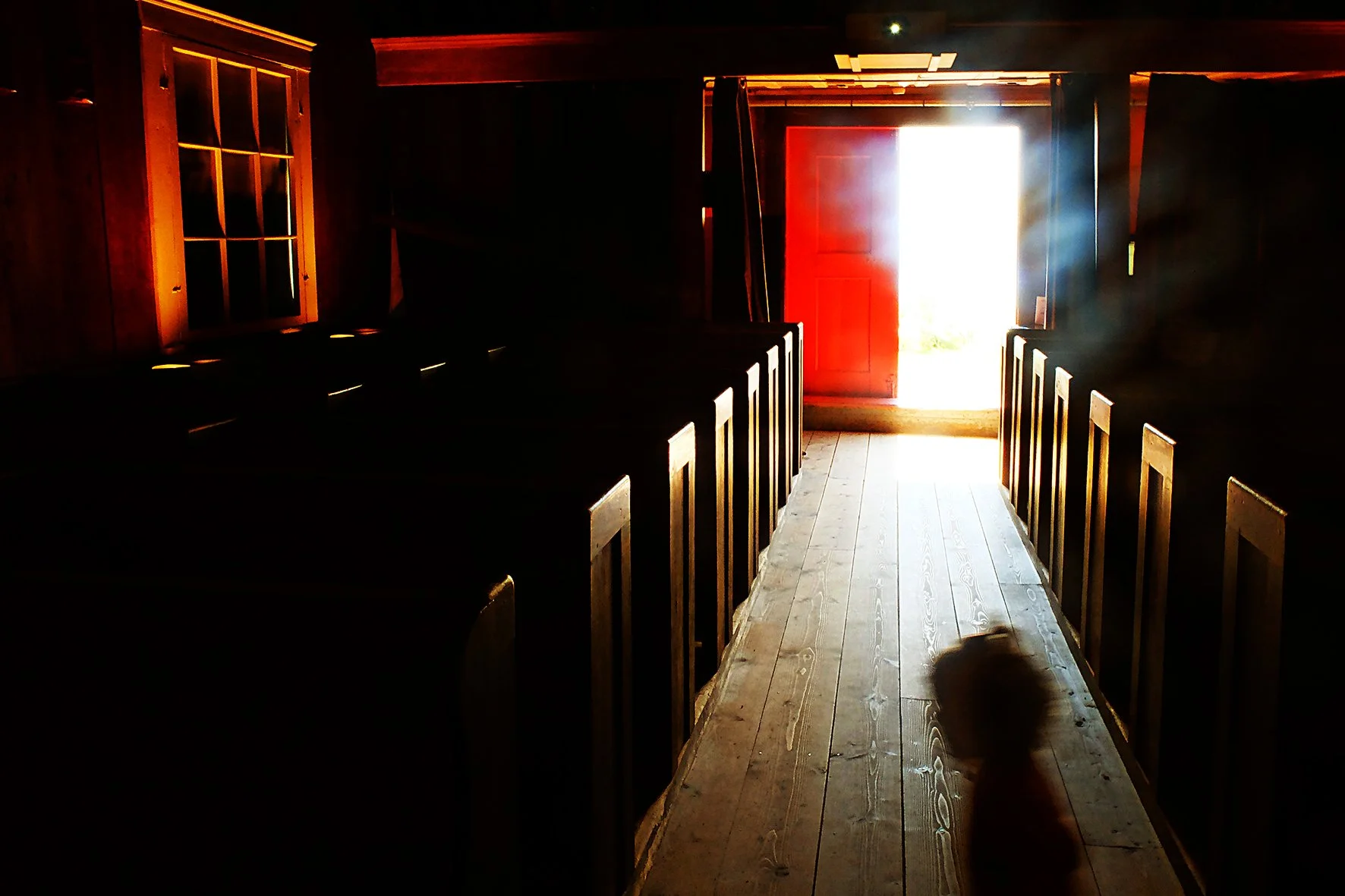 Intérieur sombre d'une église en bois, avec une porte rouge ouverte laissant entrer de la lumière, un enfant en silhouette au sol.