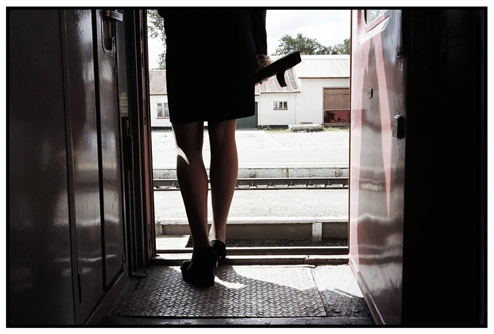 Une femme en jupe et talons hauts se tient dans la porte d'un train, tenant un journal ou un magazine, regardant dehors vers une gare avec des bâtiments en arrière-plan.