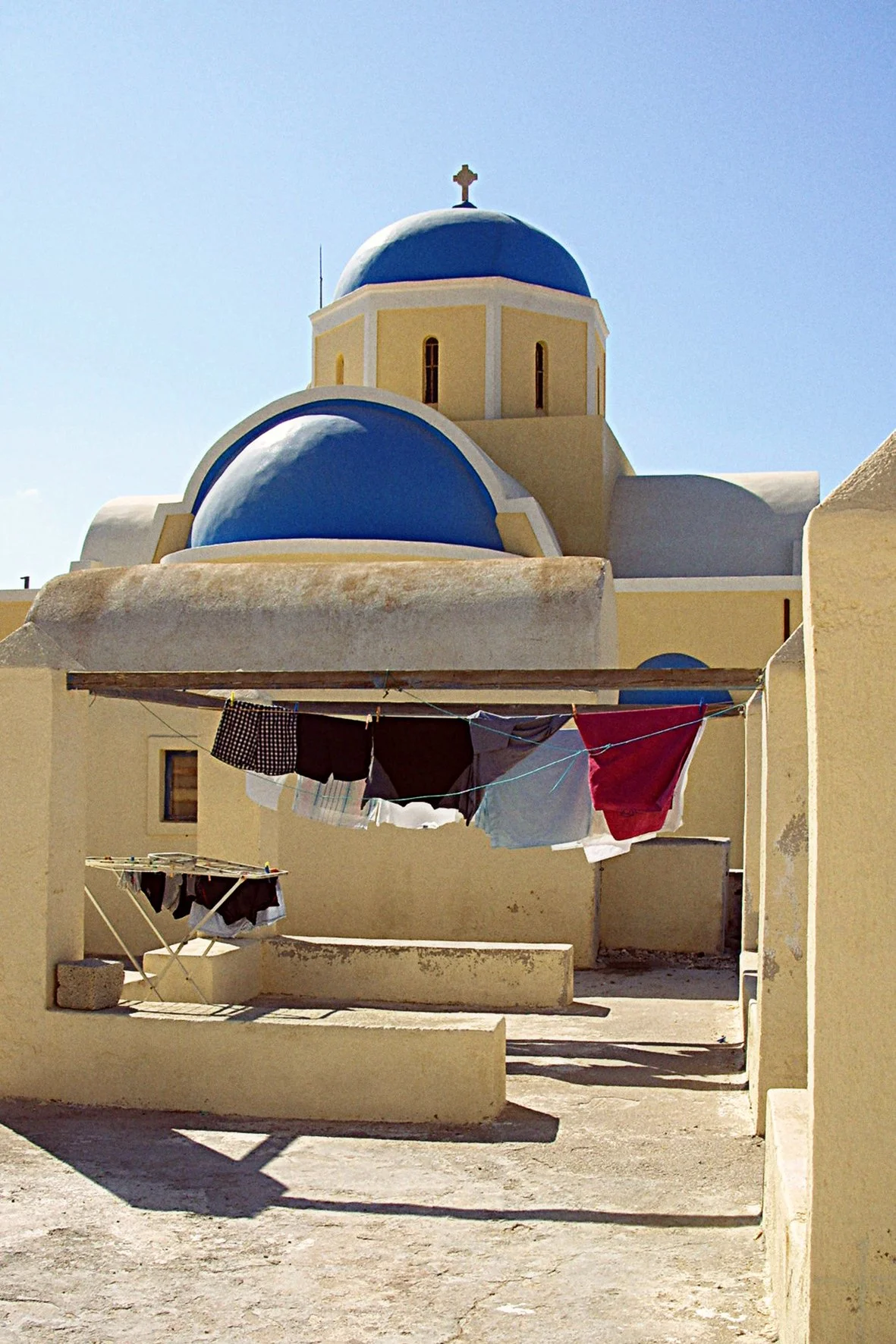 Une église avec un toit en coupole bleu et un mur beige, sous un ciel clair, avec des vêtements suspendus à une corde à linge dans la cour.