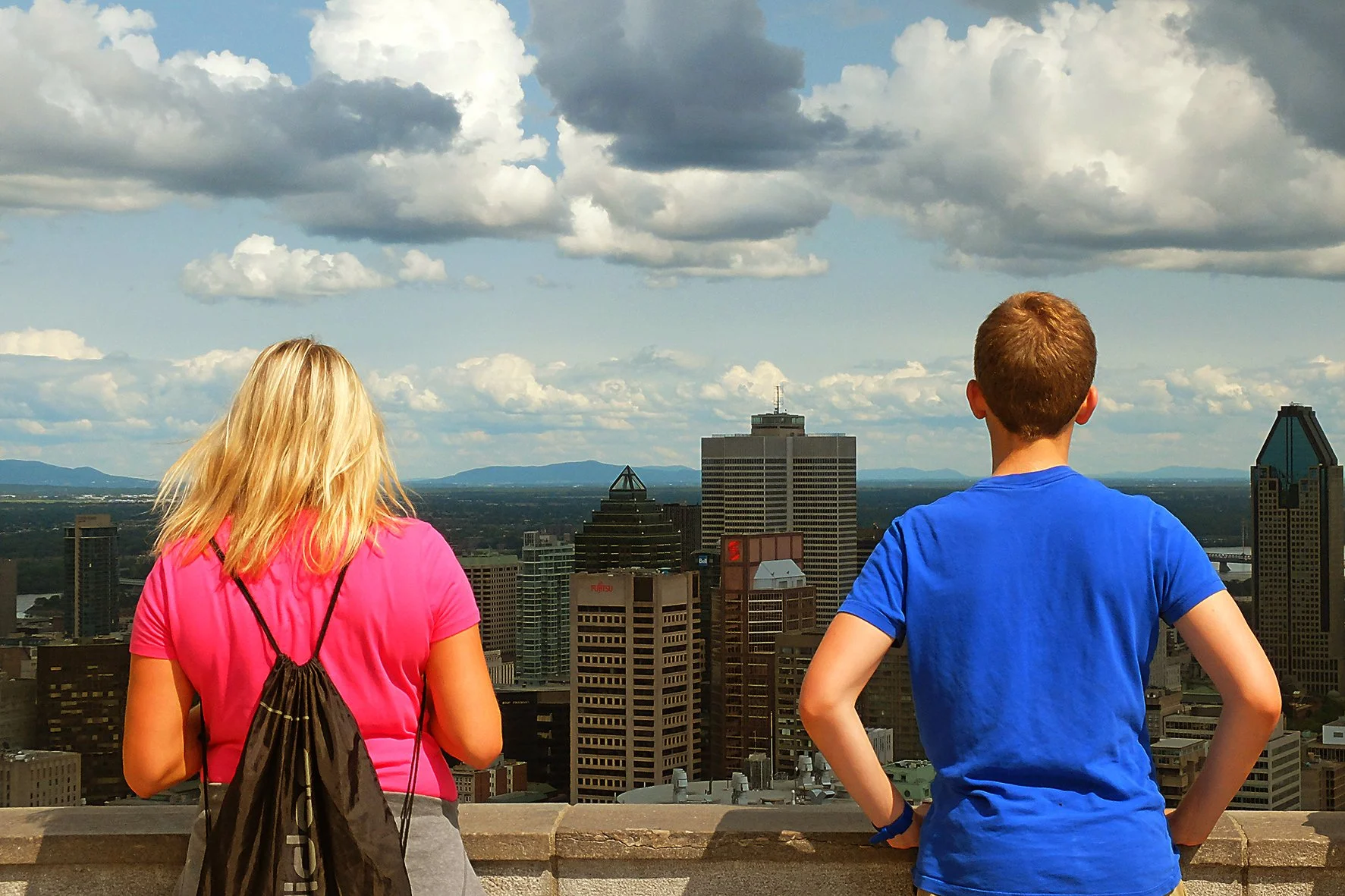 Deux jeunes regardent la ville depuis un point de vue élevé, sous un ciel partiellement nuageux.