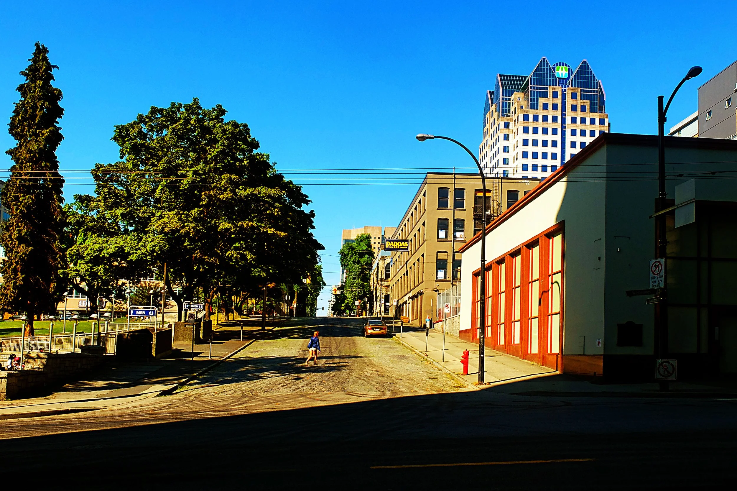 Une rue urbaine avec des arbres, un bâtiment beige, une personne marchant, et un gratte-ciel en arrière-plan.
