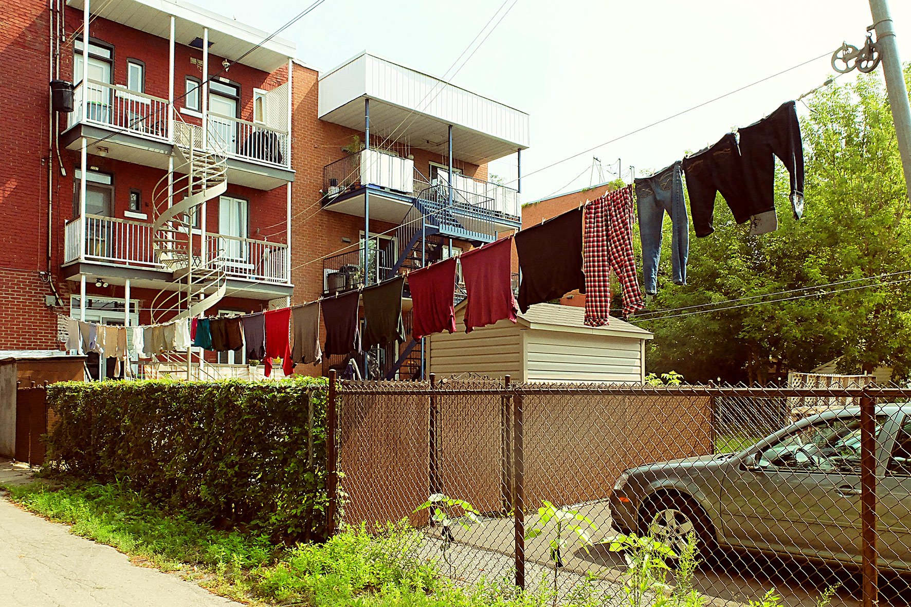 Lave-linge accrochant des vêtements suspendus à une ligne de lavage dans une cour arrière résidentielle, avec un bâtiment en brique en arrière-plan, un arbre verdoyant et une voiture garée derrière la clôture.