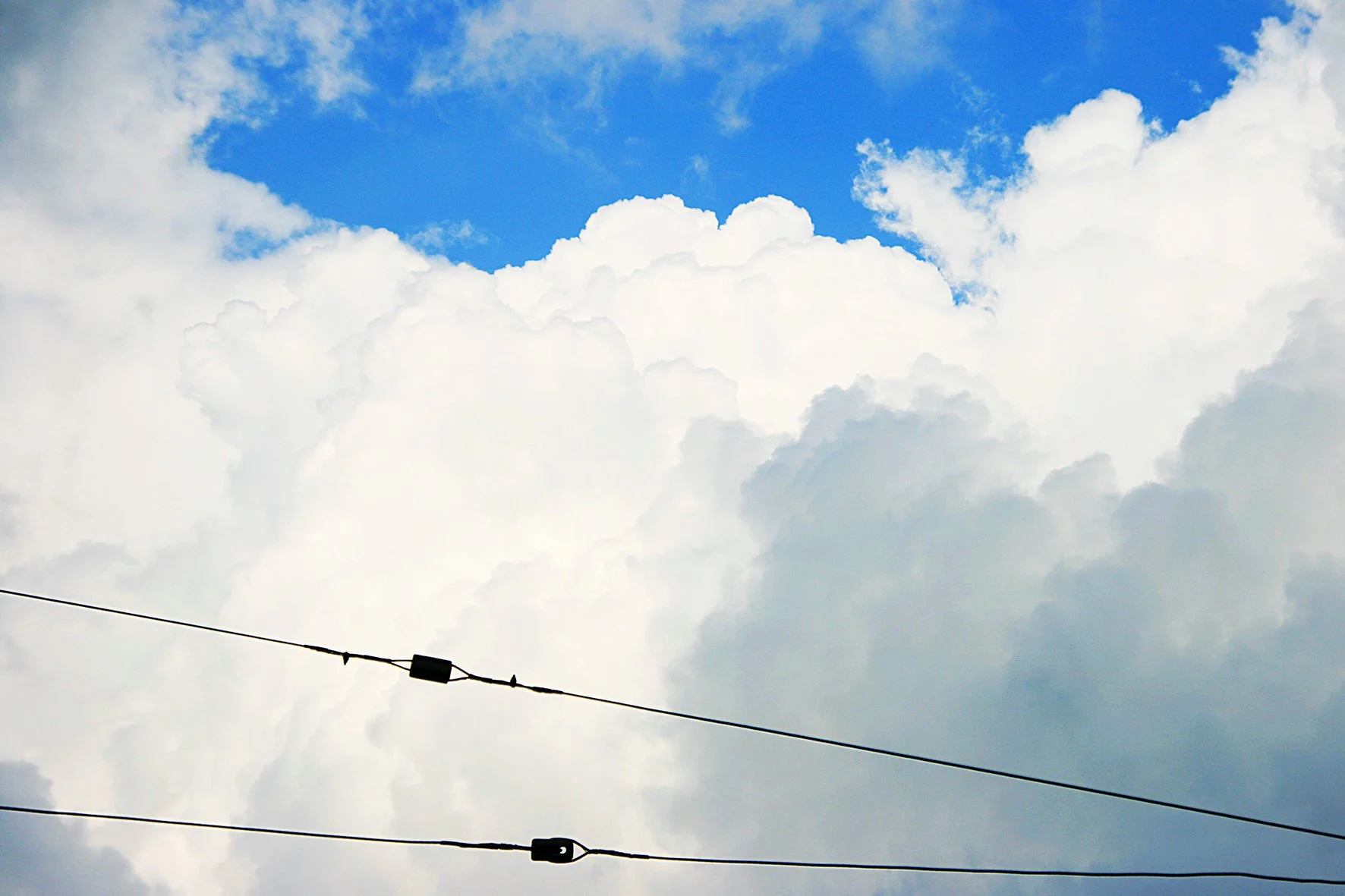 Ciel bleu avec des nuages blancs, deux câbles électriques traversent l'image horizontalement.
