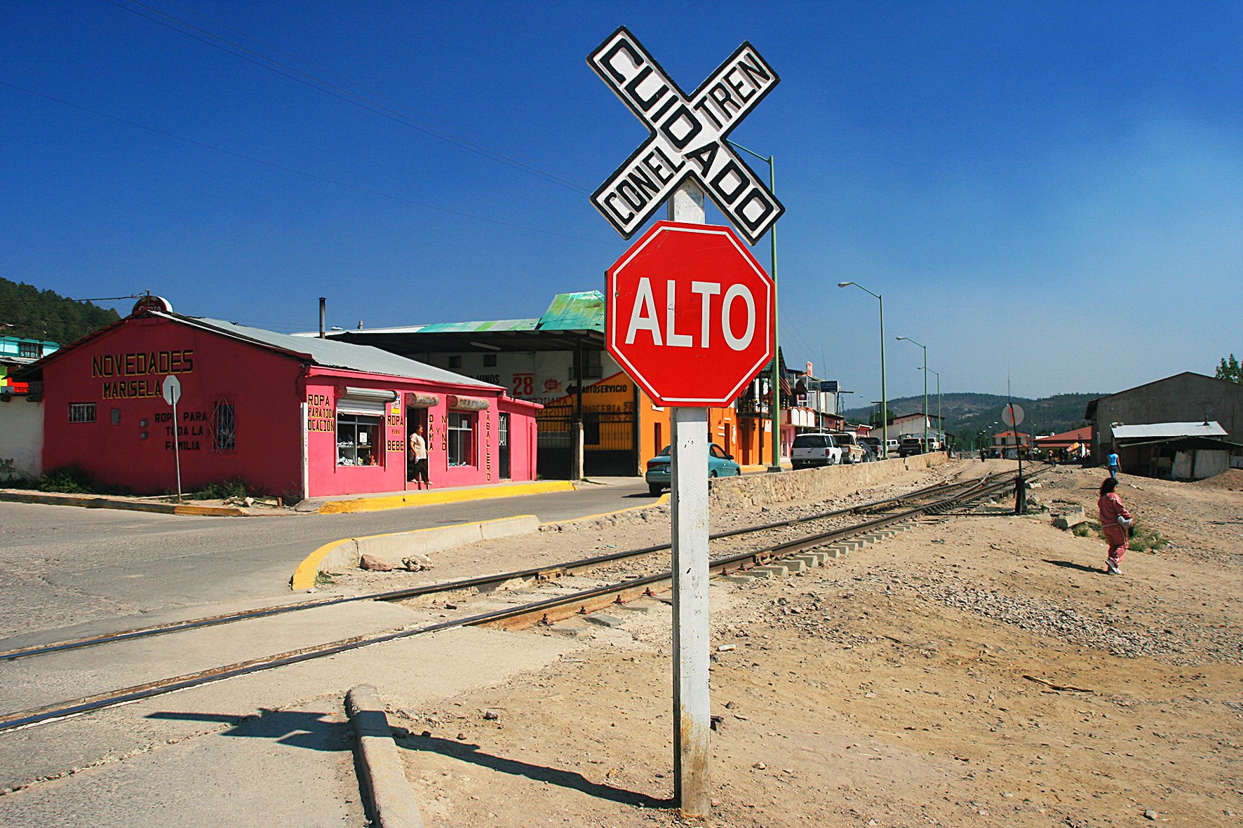 Un panneau d'arrêt rouge avec inscription 'ALTO' et un panneau de niveau de croisement de chemin avec l'inscription 'CONEL TRAM'. En arrière-plan, des bâtiments colorés, dont un rose, et des voies ferrées dans un environnement de petite ville en zone