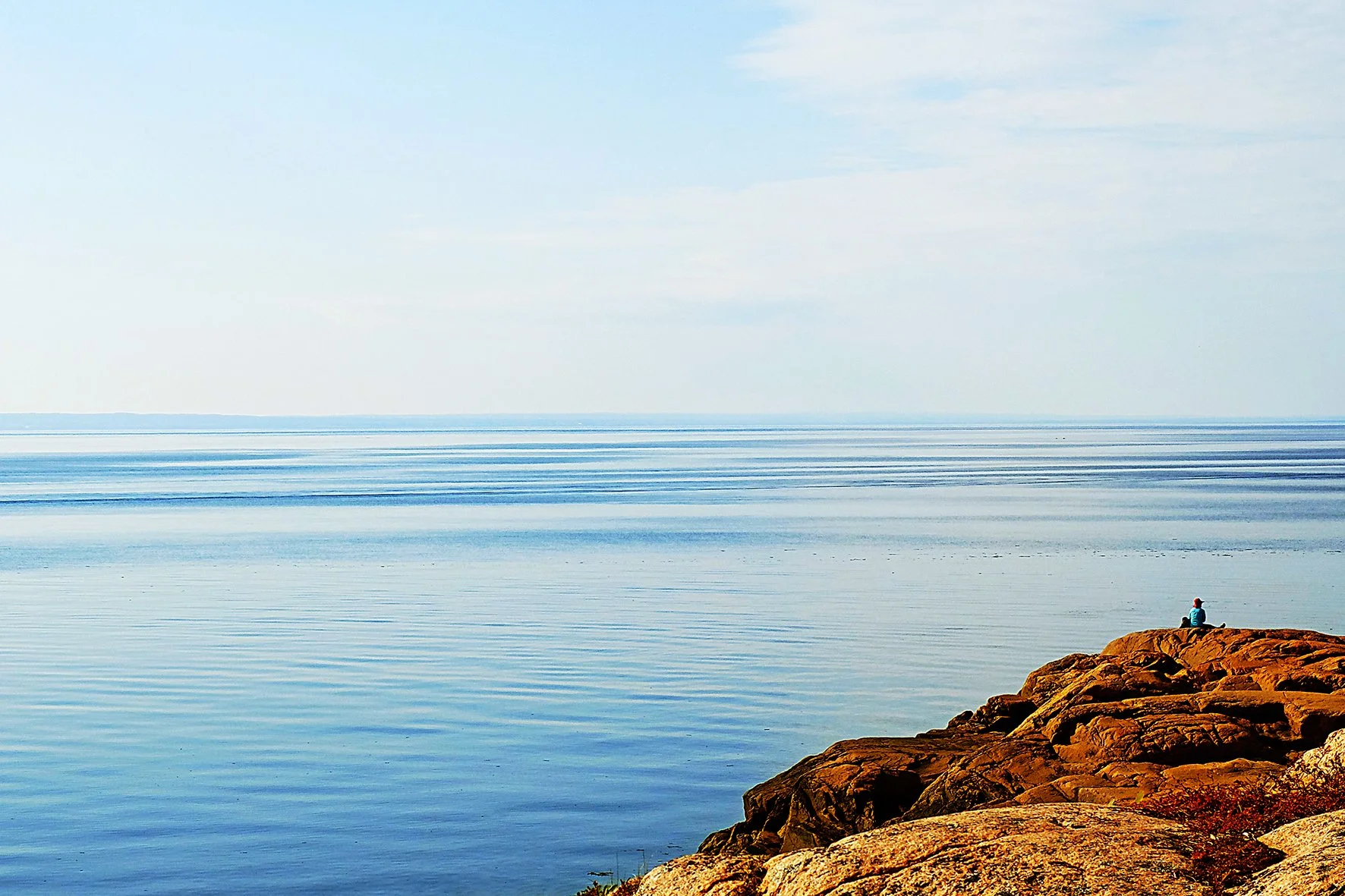 Vue d'un lac ou d'une mer calme avec un rocher en premier plan et une personne assise au bord de l'eau regardant l'horizon.