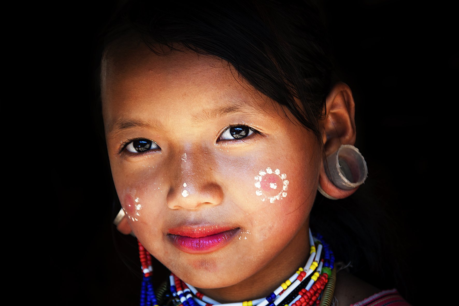 Jeune fille souriante avec tresses, maquillage traditionnel et bijoux colorés, portant des décorations blanches sur les joues, avec une peau tannée, sur fond noir.