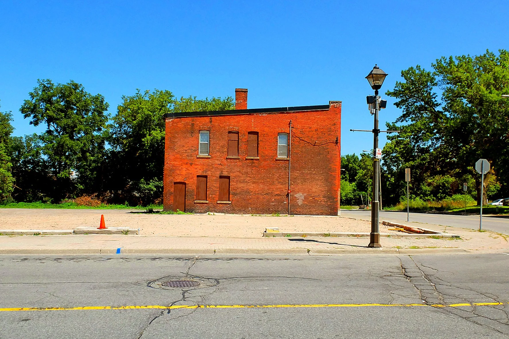Bâtiment en brick rouge abandonné avec des fenêtres et une porte barricadées, verdure à l'arrière-plan, lampadaire sur le trottoir, route en premier plan, ciel bleu clair.
