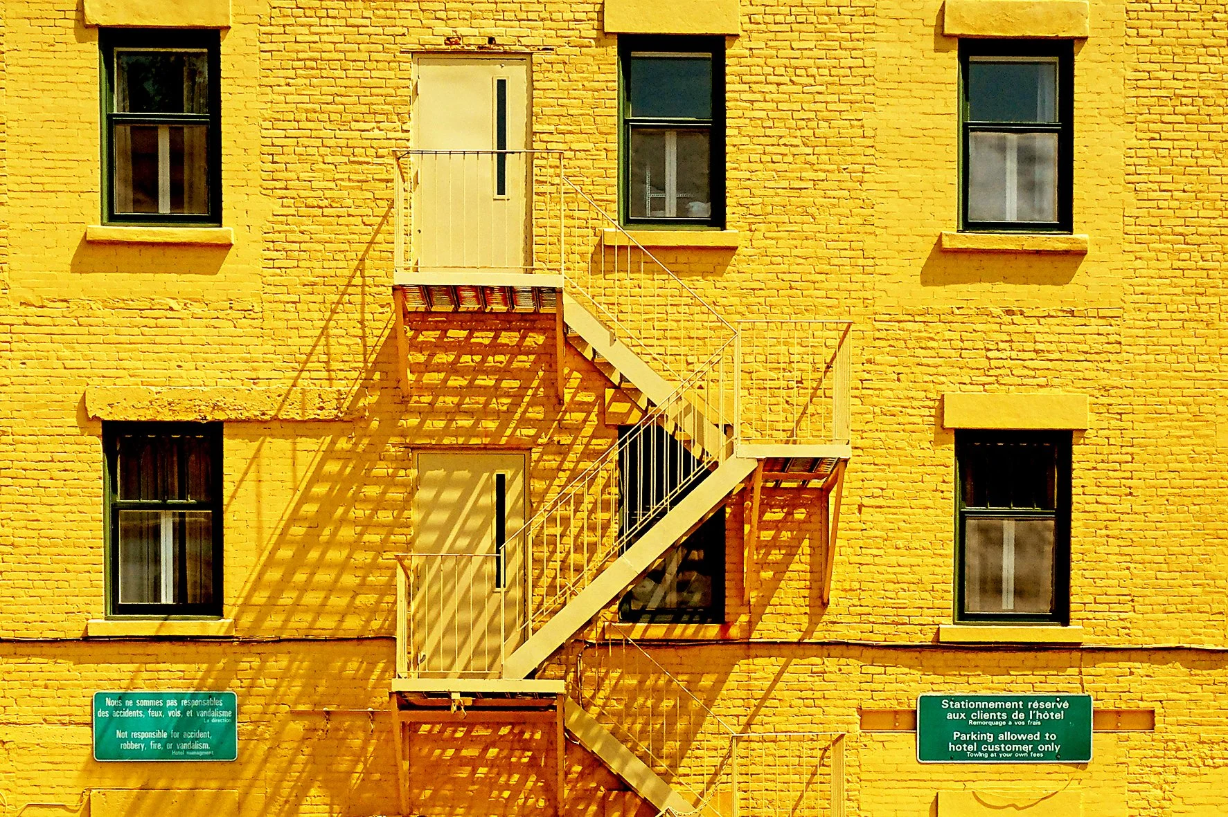 Façade d'un bâtiment jaune avec une échelle de secours blanche et deux panneaux en bas, un en français et un en anglais. Quatre fenêtres visibles.
