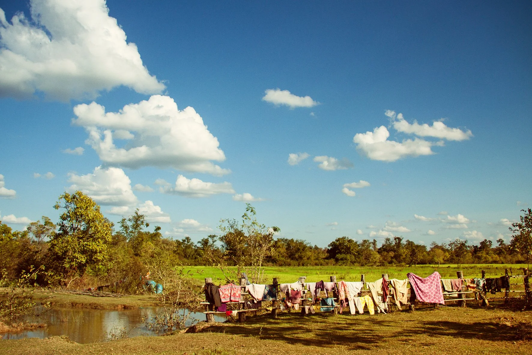 Le paysage rural montre un ciel bleu avec des nuages blancs, un champ vert avec des arbres, et un étang. Sur la clôture en bois, des vêtements et des tissus sont suspendus pour sécher.