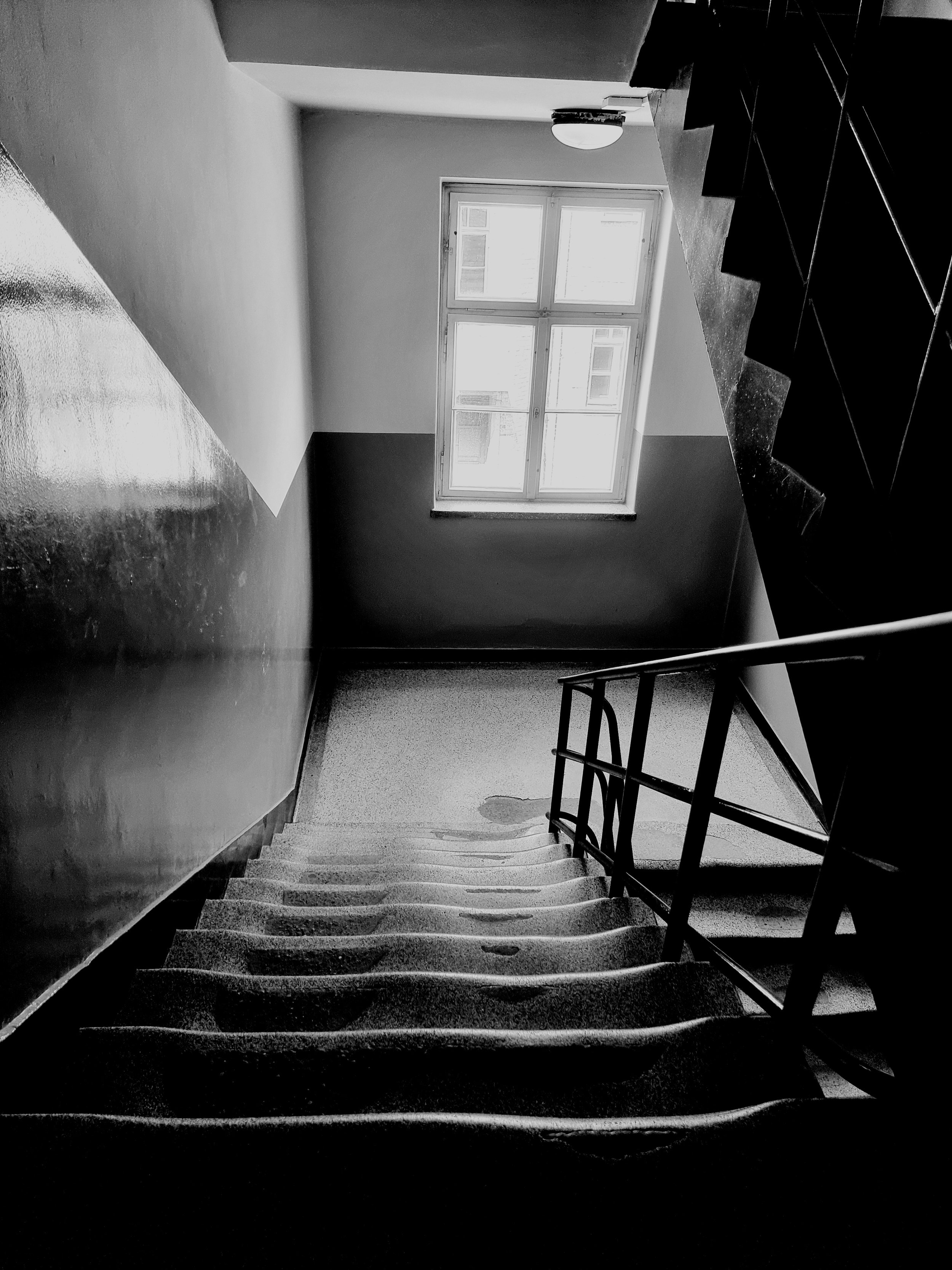 Escalier en béton avec rampe métallique, vue du haut vers le bas, dans un bâtiment intérieur, avec une fenêtre lumineuse au fond.