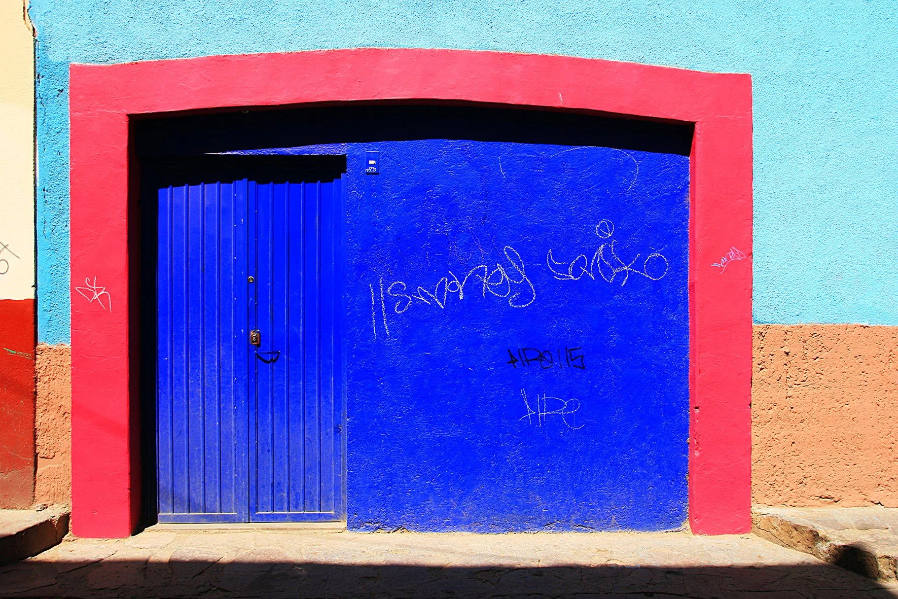 Porte bleue avec un encadrements rose, graffitis en blanc et noir sur le mur bleu, façade colorée avec des sections en turquoise, rose et beige
